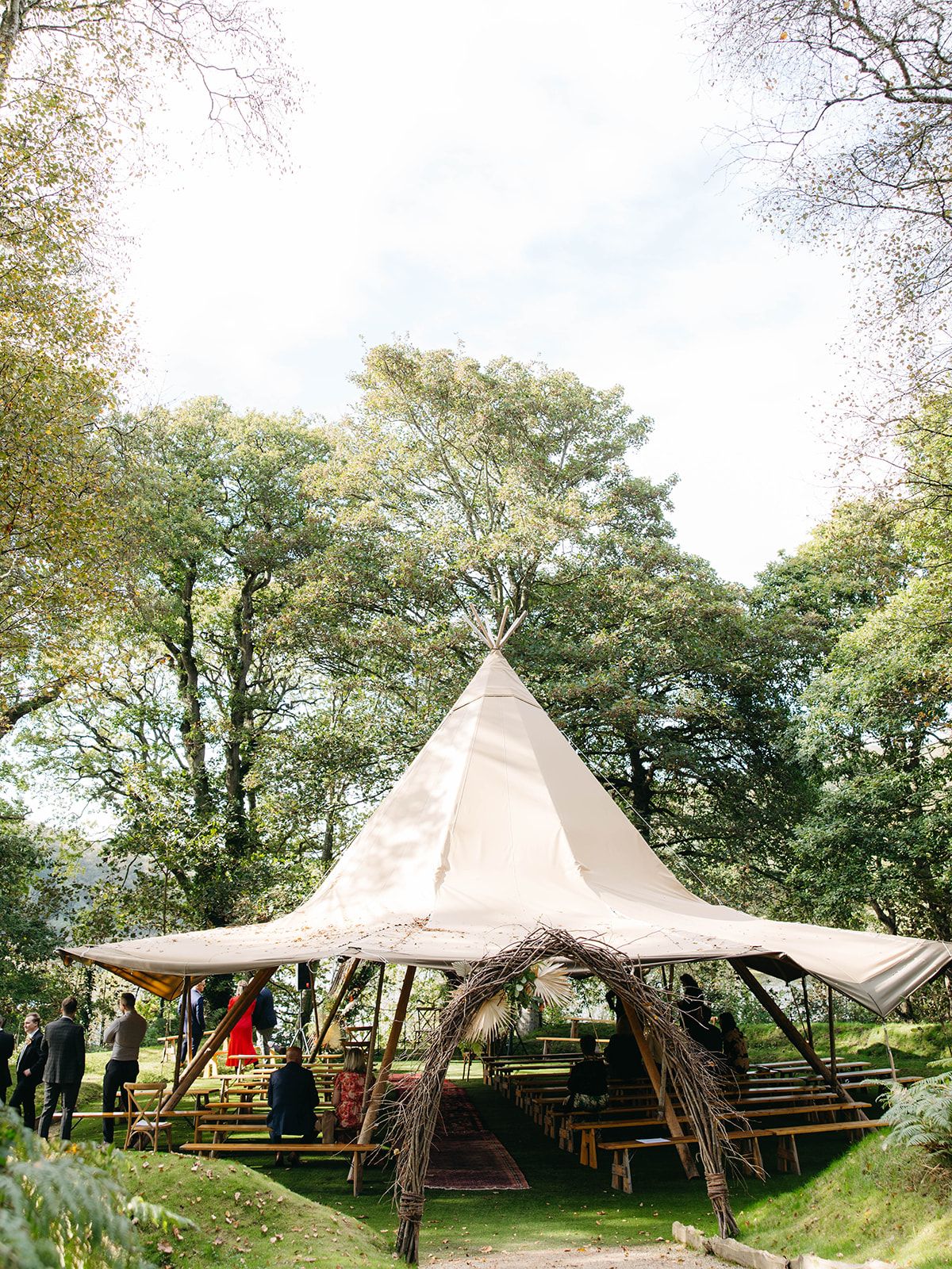 A rustic teepee-style wedding tent with wooden benches set up in a lush green forest setting.