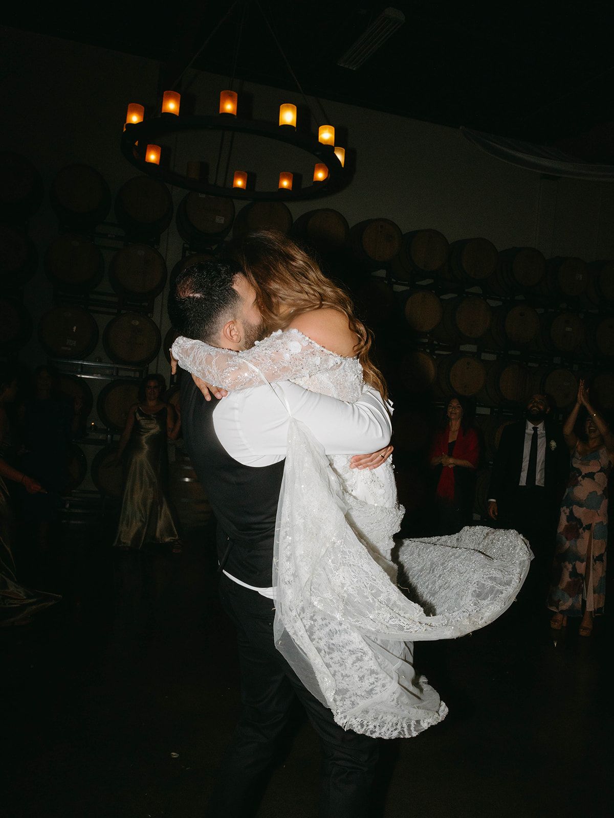 A couple embraces during their first dance in a rustic winery cellar with romantic barrel decor and overhead lighting.