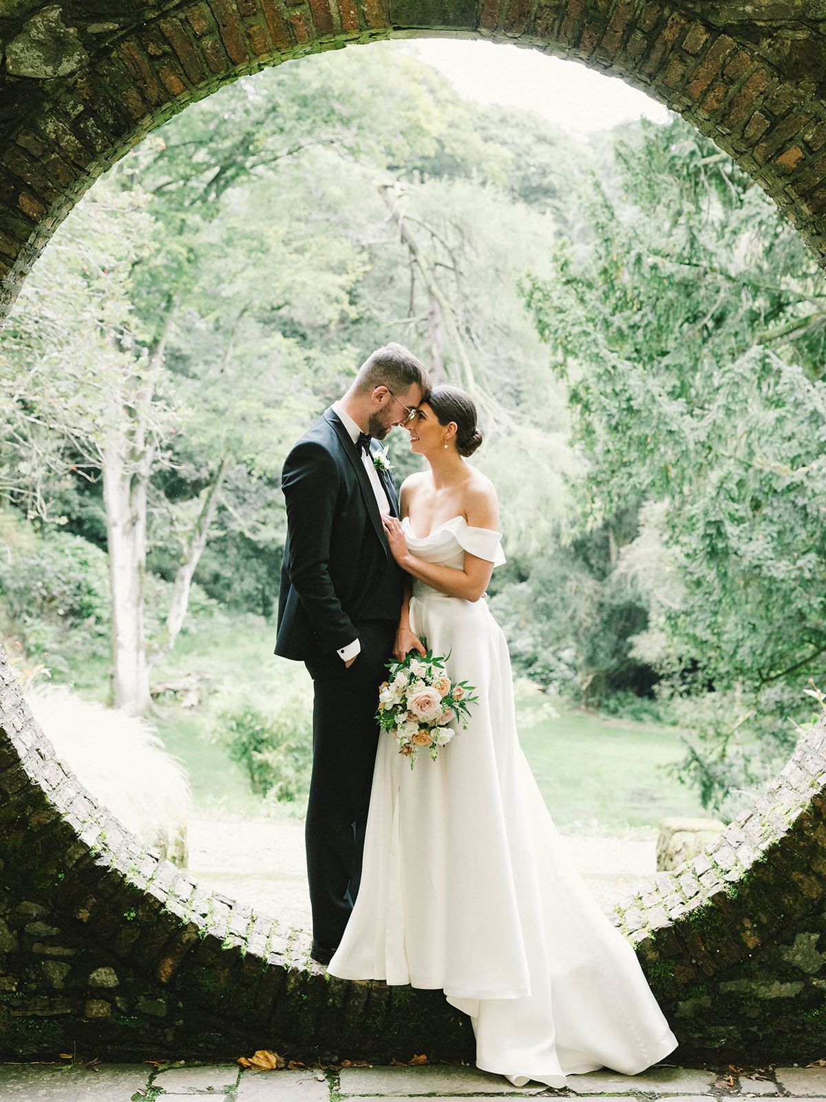 A loving pair embraces beneath a round stone arch surrounded by dreamy gardens and foliage.