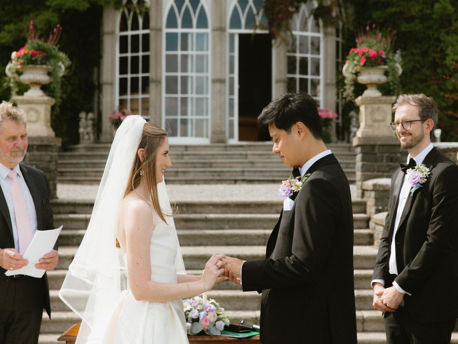 Wedding ceremony on stone steps with Gothic windows and floral decorations in background.