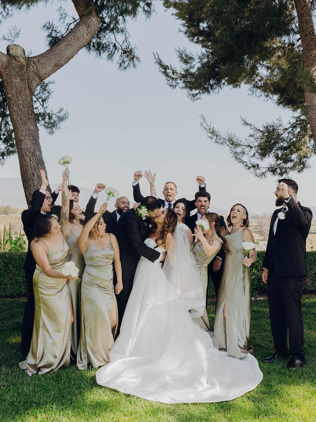 Wedding party celebrates outdoors under a large tree with champagne while wearing pale gold and black formal attire.