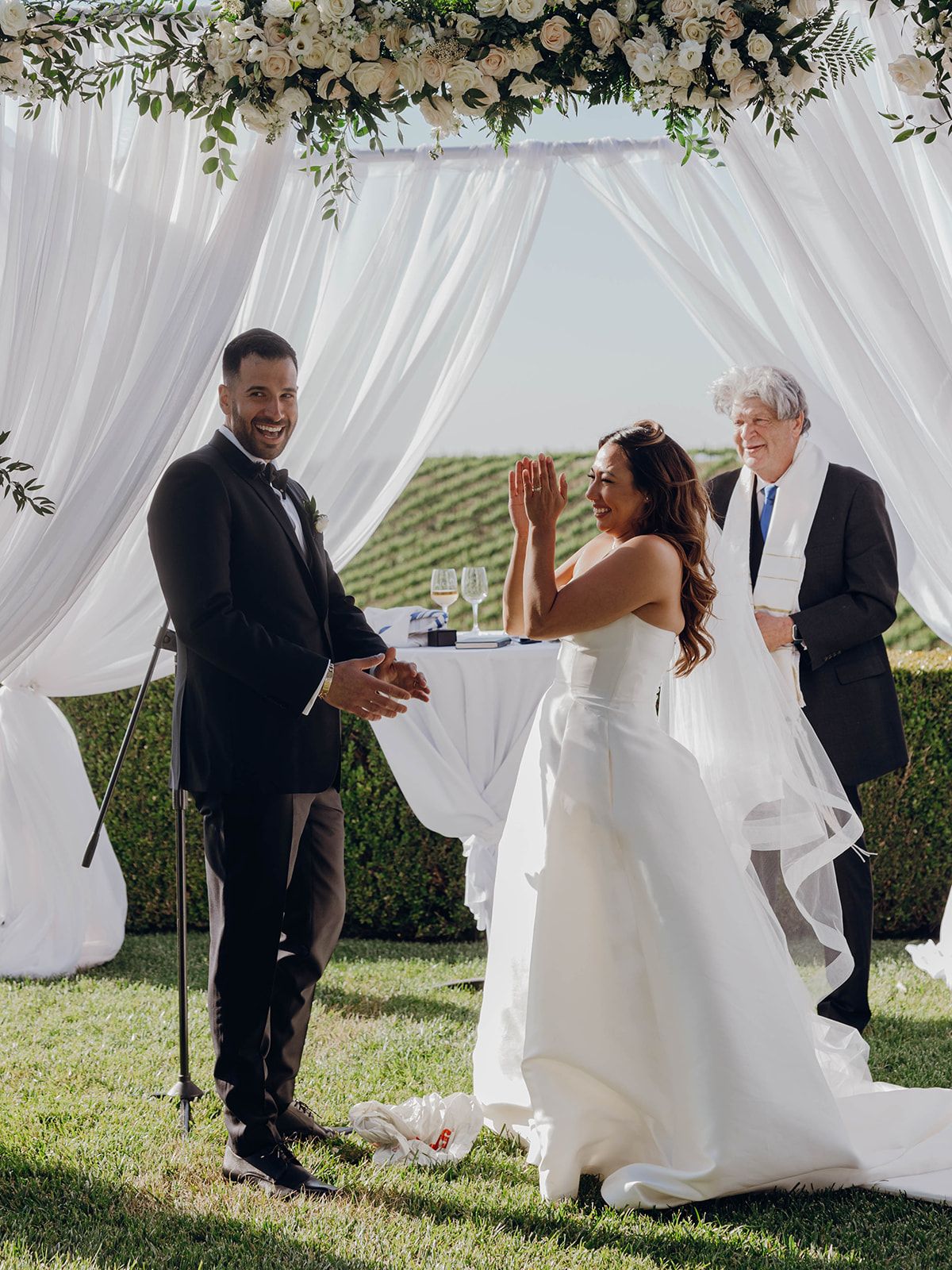 A joyful outdoor wedding ceremony under a white draped arch decorated with greenery in a vineyard setting.