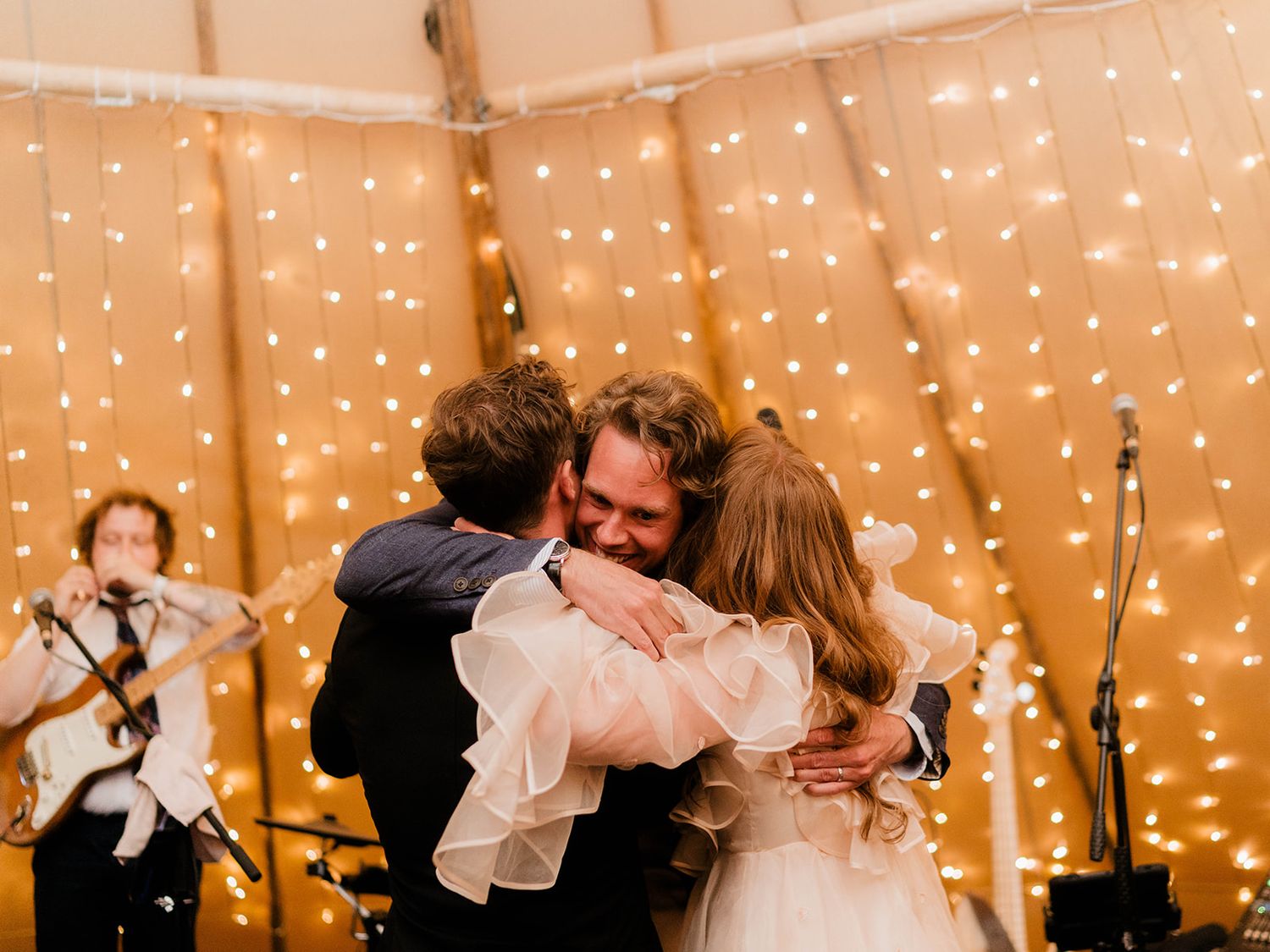 Emotional group hug during wedding reception with string lights in background.