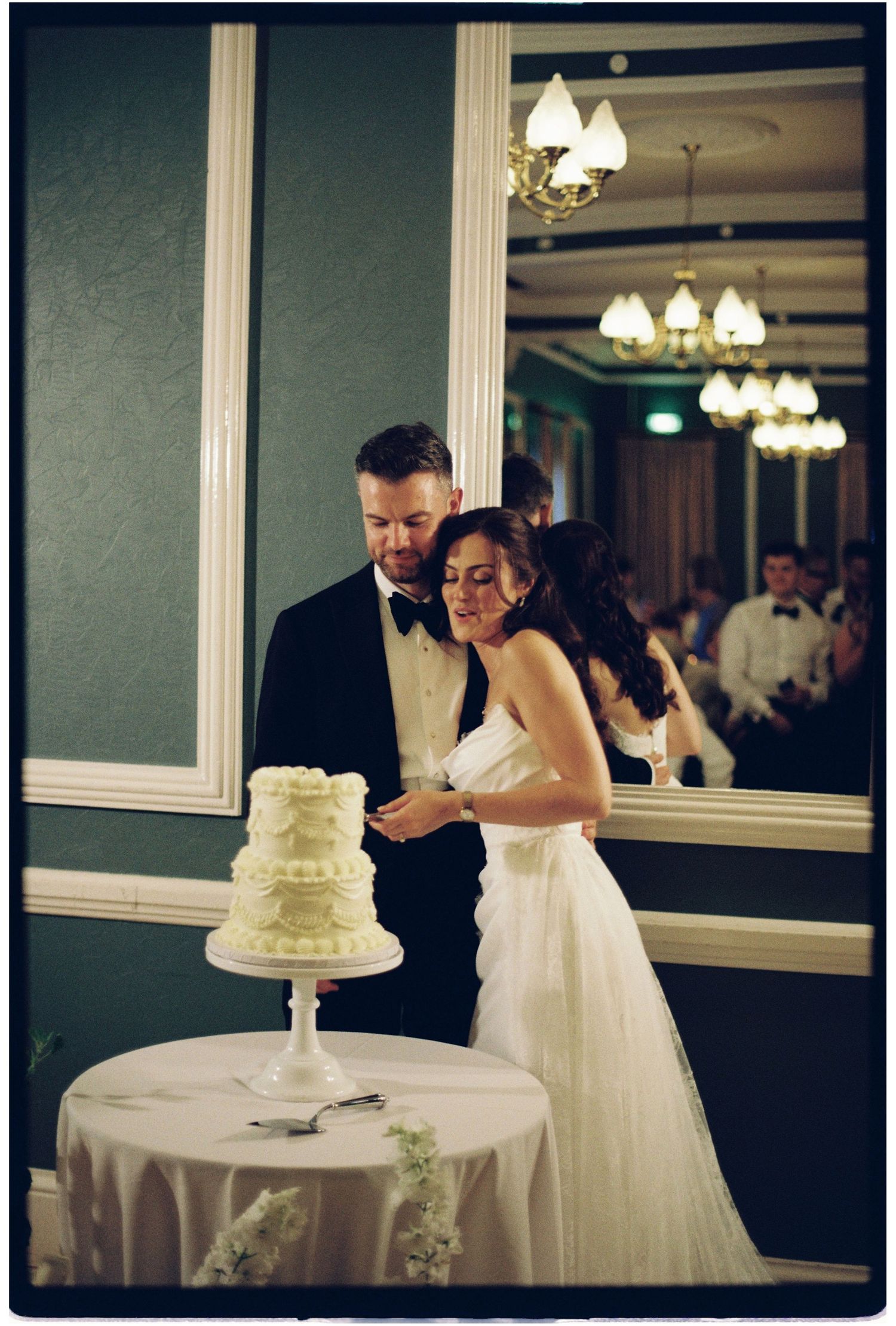Bride and groom cutting their white tiered wedding cake at elegant reception venue with chandelier reflection in mirror.