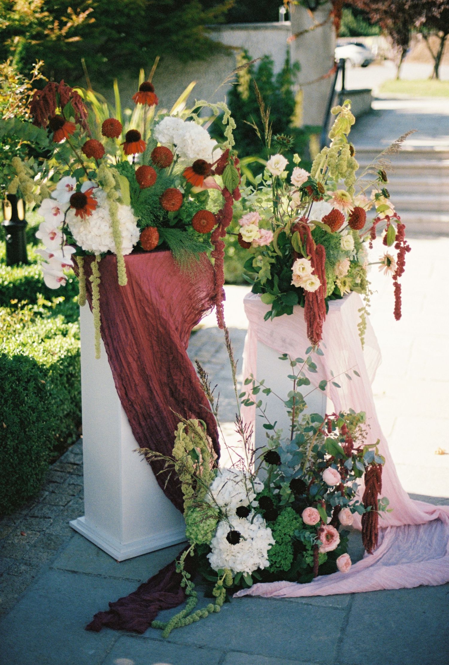 Elegant wedding ceremony decor with burgundy draped fabric, white pedestals, and lush floral arrangements of roses and greenery.