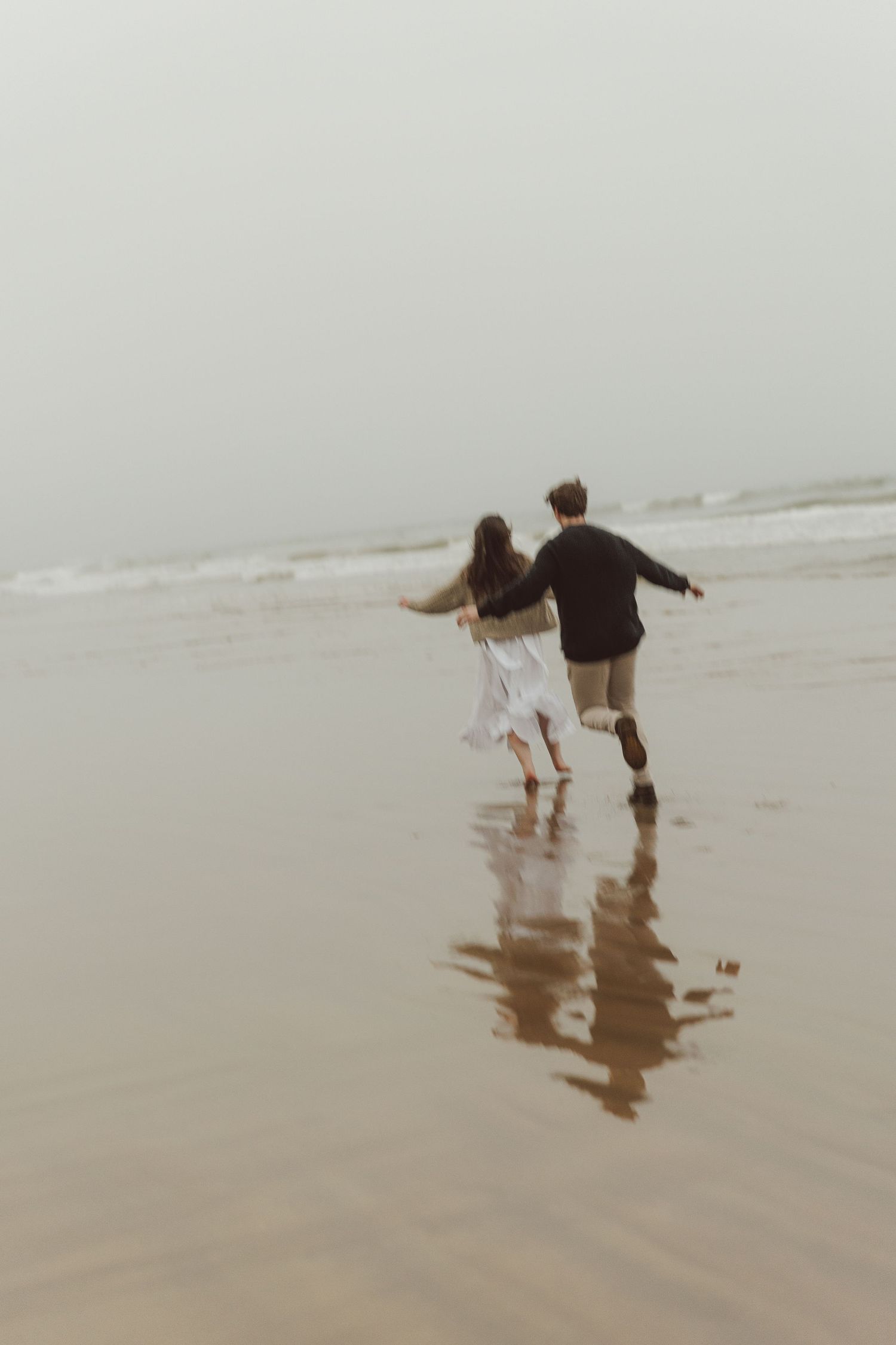Couple running hand-in-hand on a misty beach, reflecting on wet sand, woman in white dress, man in dark sweater.