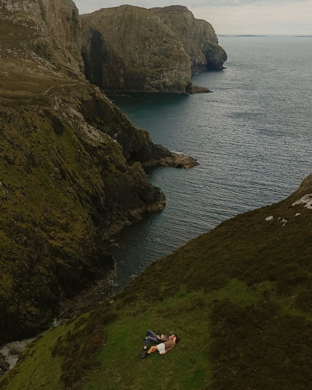 Couple lying on green cliffside overlooking dramatic rocky coastline and calm blue sea in Ireland.