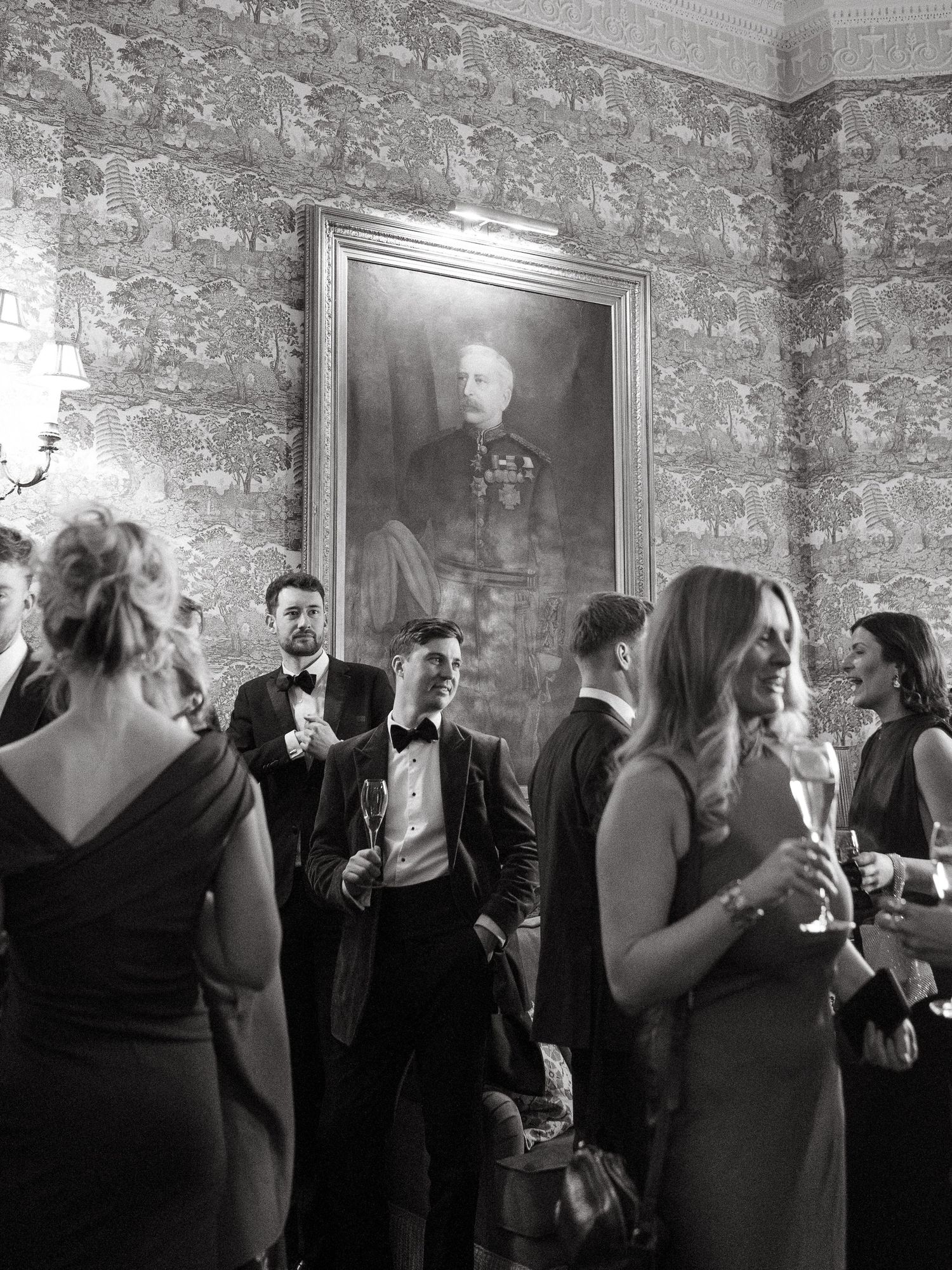 Elegant black and white wedding reception photo with guests in formal attire beneath a classic portrait painting.