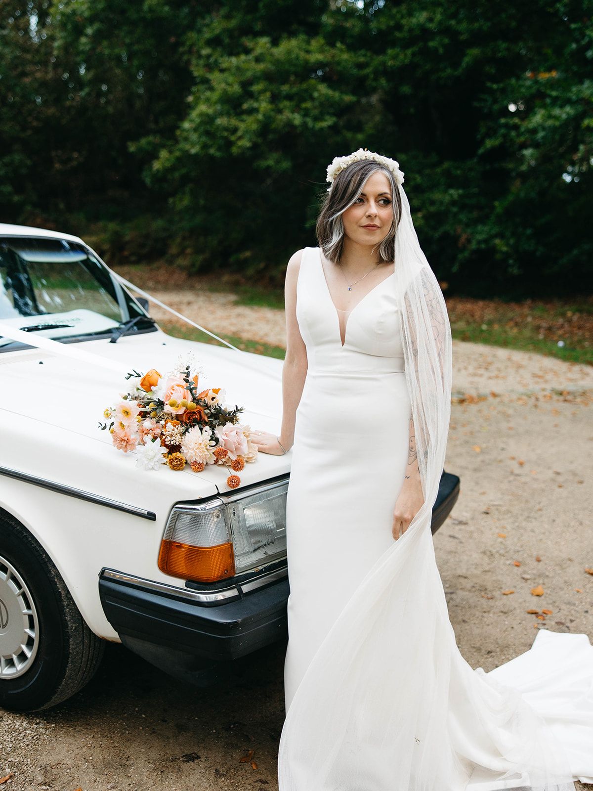 A bride in a flowing white wedding dress stands next to a classic white car decorated with autumn-colored flowers.