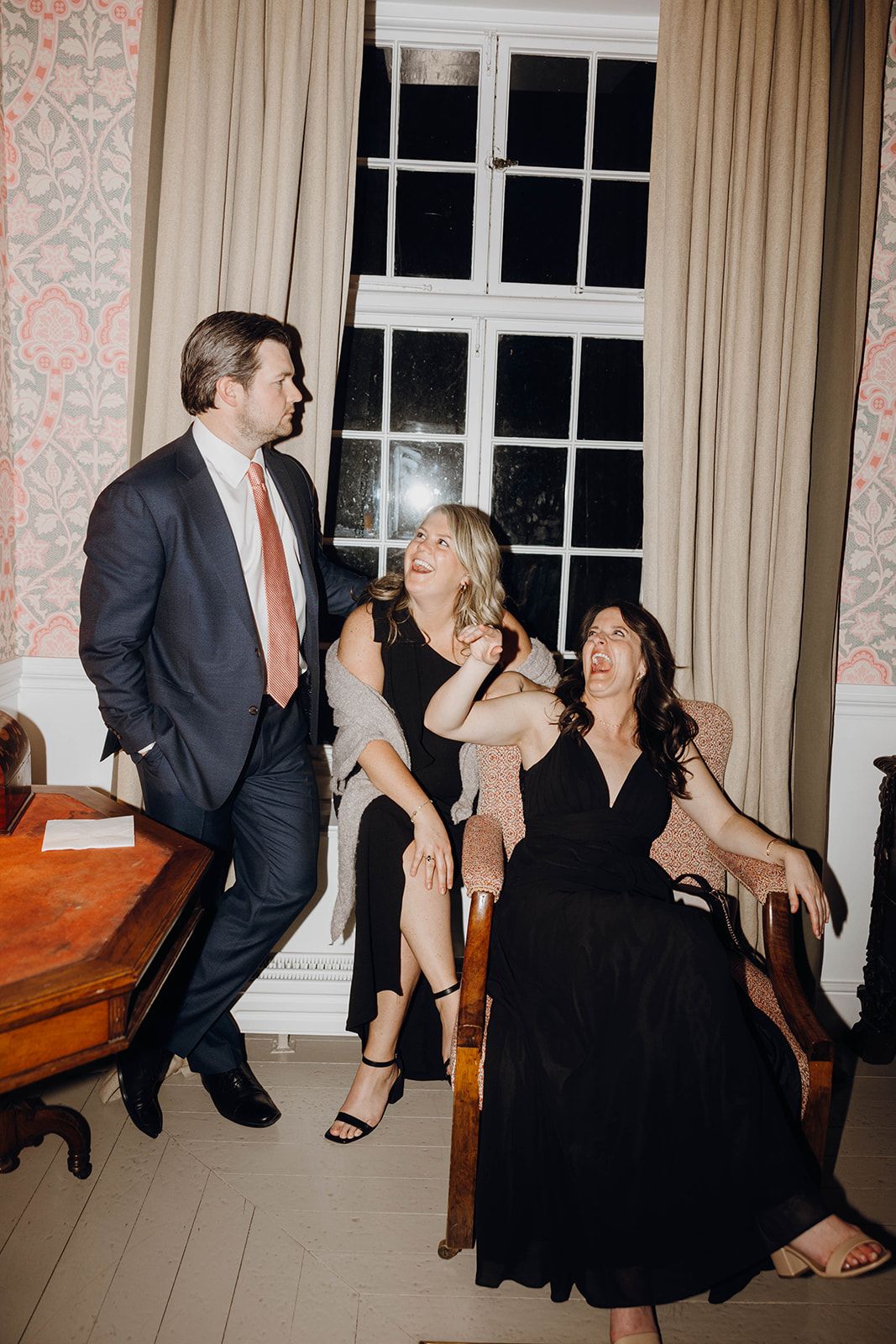 Three people sharing a candid moment of laughter at an indoor event with window and curtains in the background.