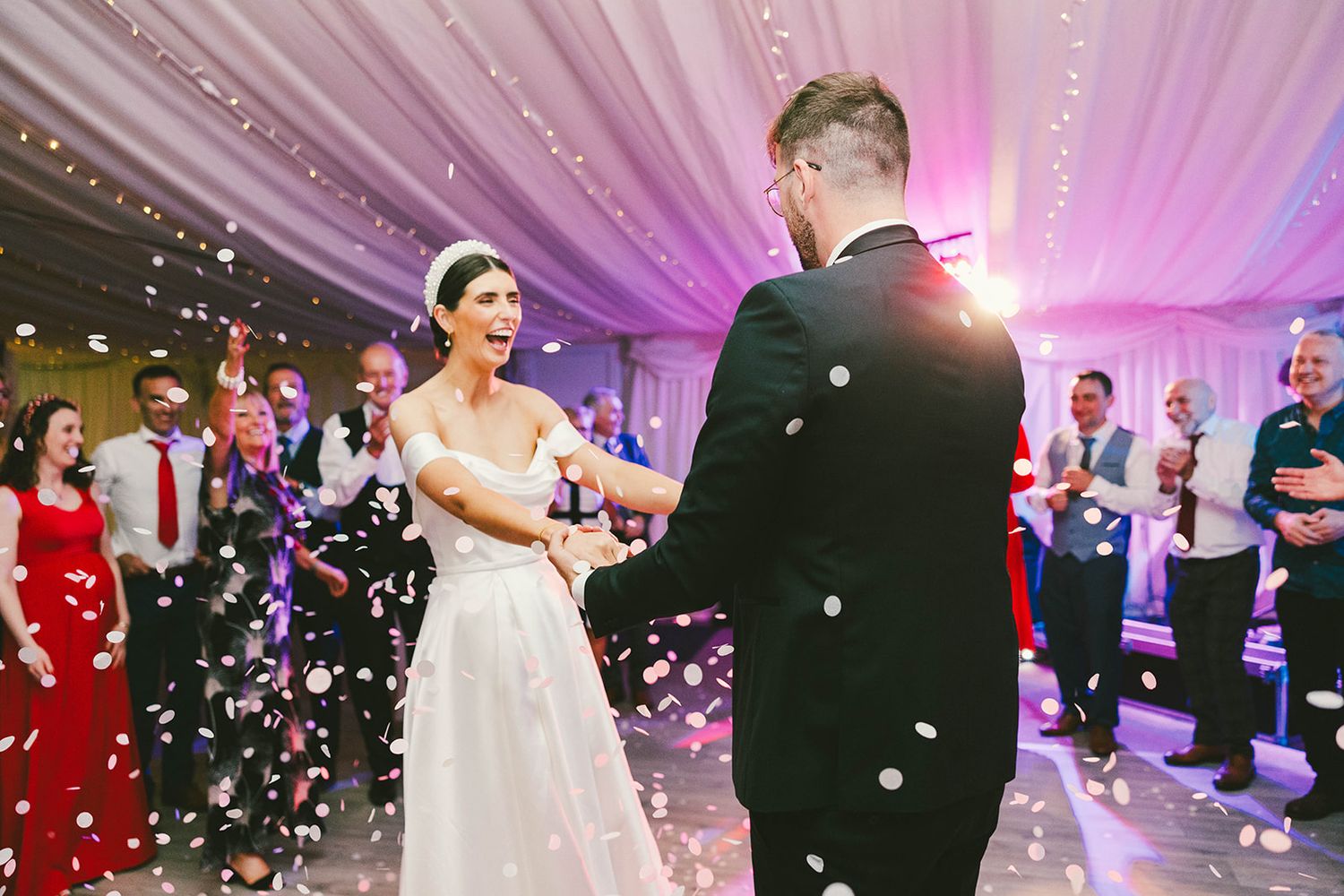 A bride and groom share their first dance under purple lighting as guests throw confetti at an elegant wedding reception.