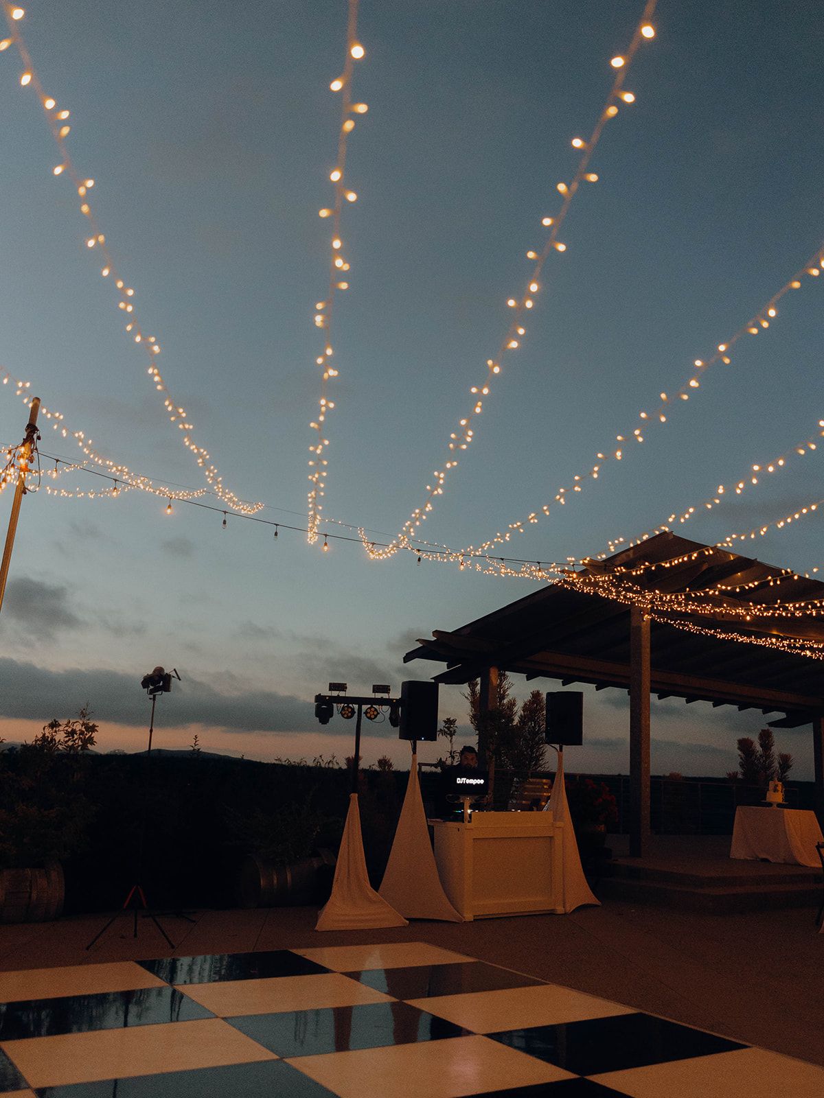 String lights illuminate an outdoor dance floor and event setup at dusk with DJ booth and checkerboard floor pattern.