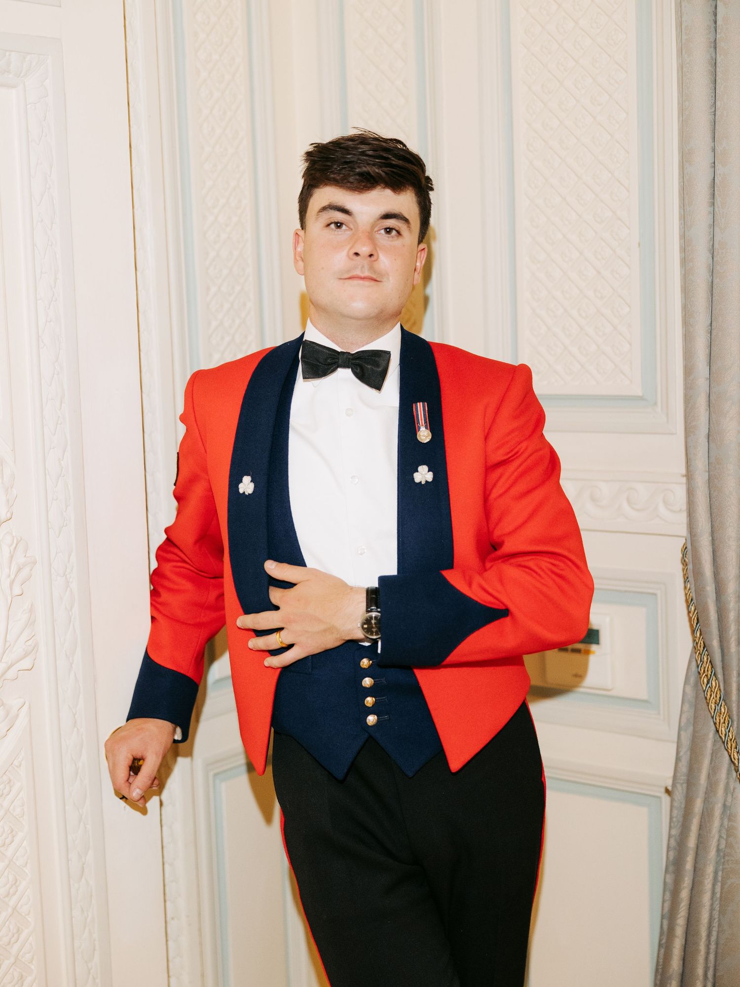 Someone in a formal red tuxedo jacket with black lapels and bow tie poses against a cream colored backdrop.