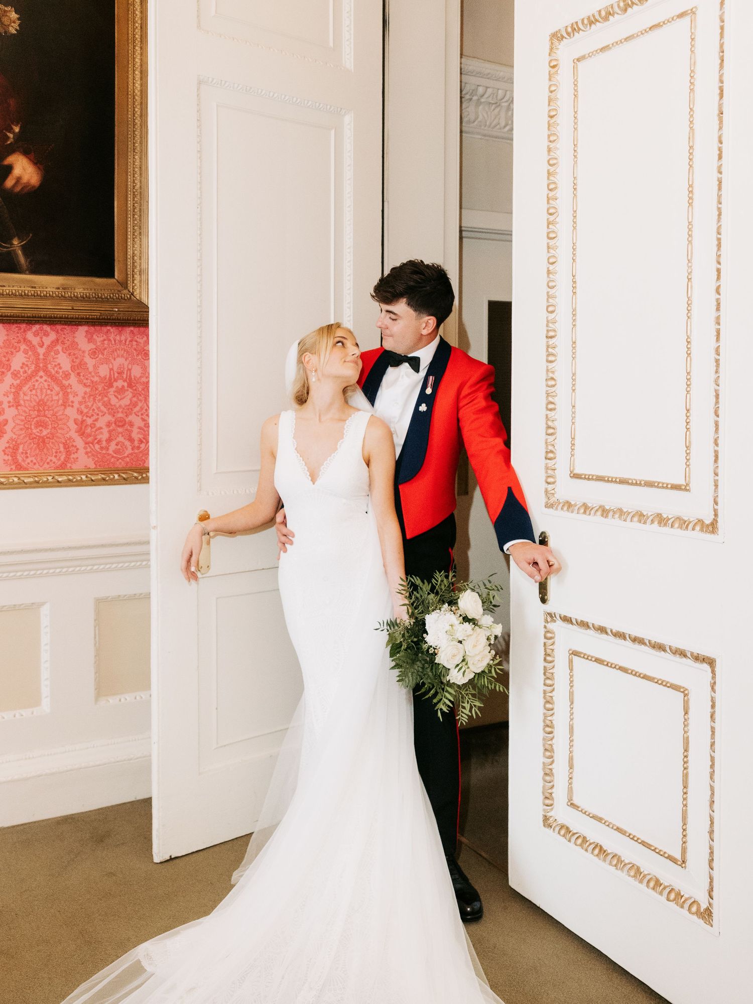 Bride in white wedding dress playfully peeks around large white door panels decorated with gold trim details.
