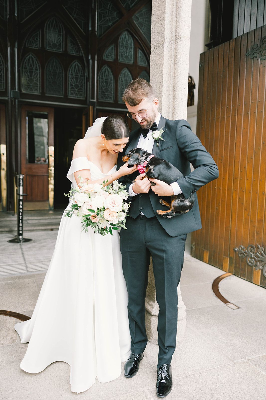 Wedding couple shares a tender moment outside a church while holding their small dog in their arms.