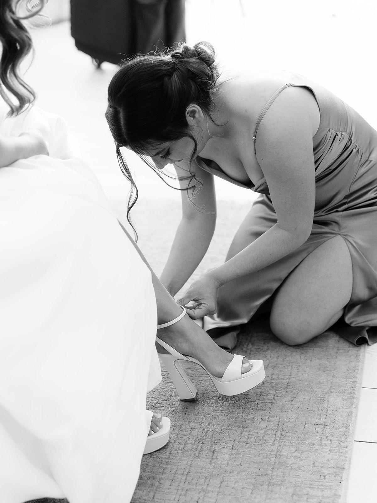 A person wearing a gray dress helps with wedding preparations, fastening a delicate white shoe on the floor.