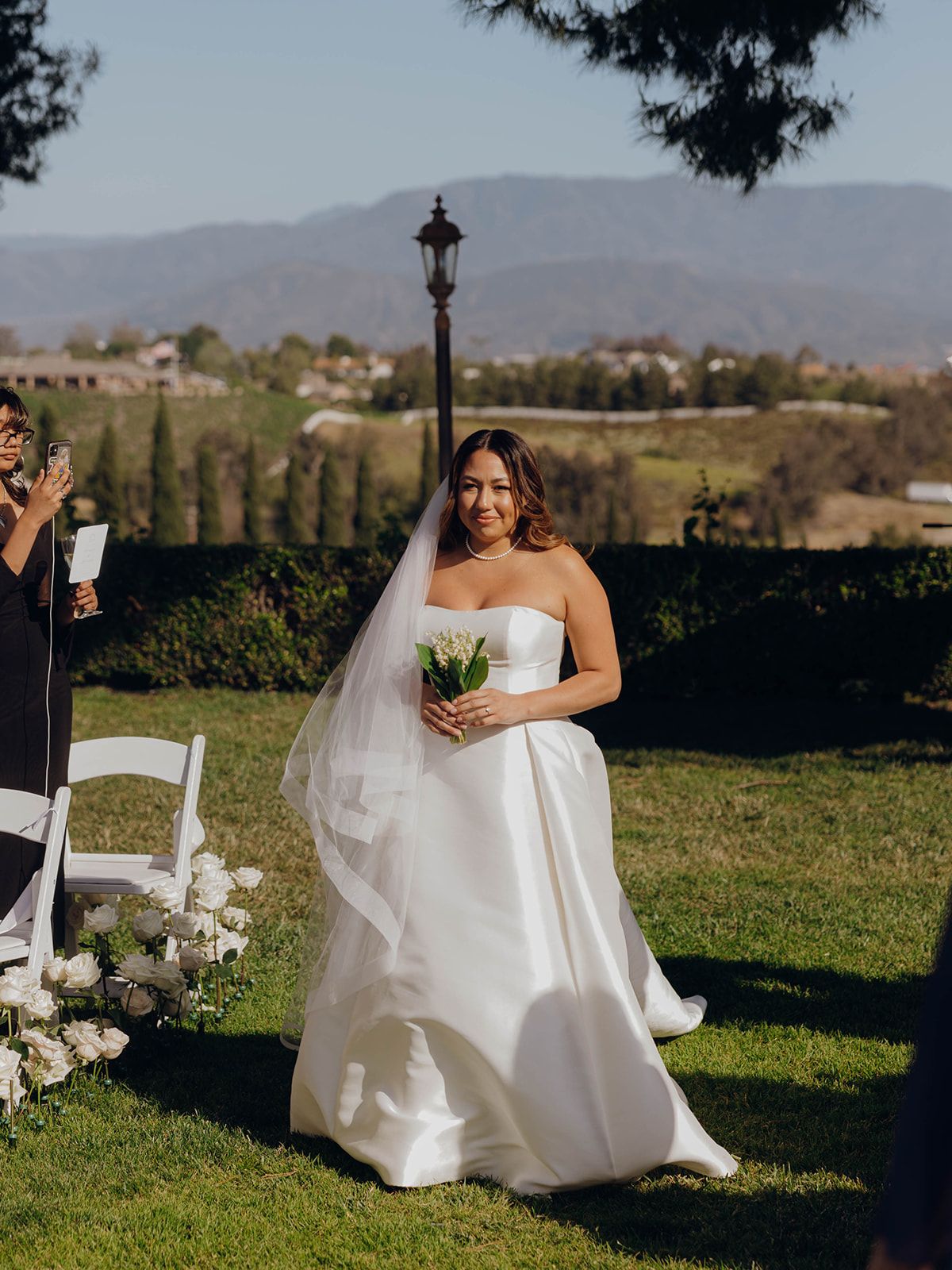 A bride in a classic white strapless wedding gown stands on a hilltop lawn with mountains visible in the background.