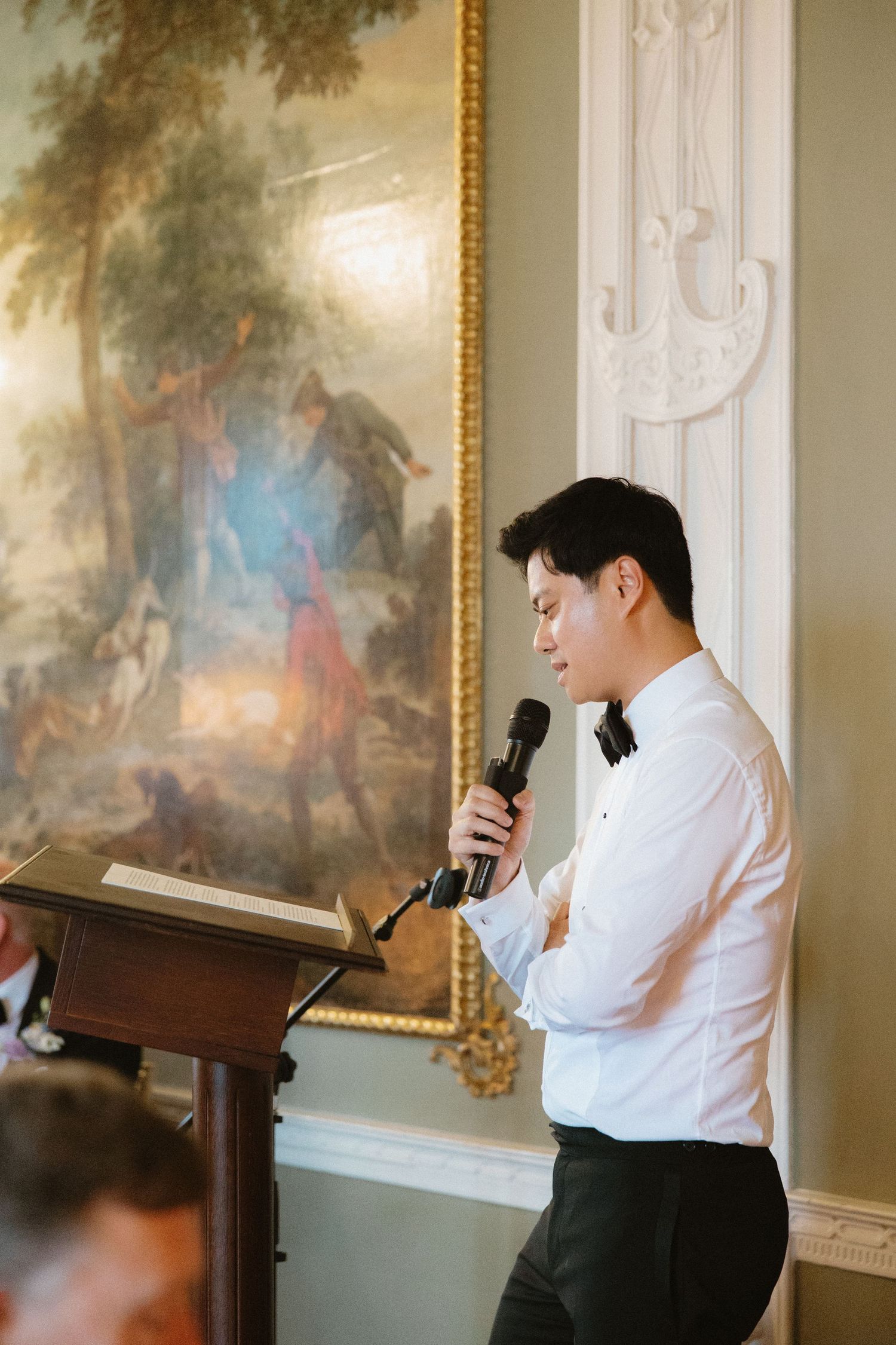 A speaker in formal attire gives a speech at an elegant venue with a classical painting in the background.