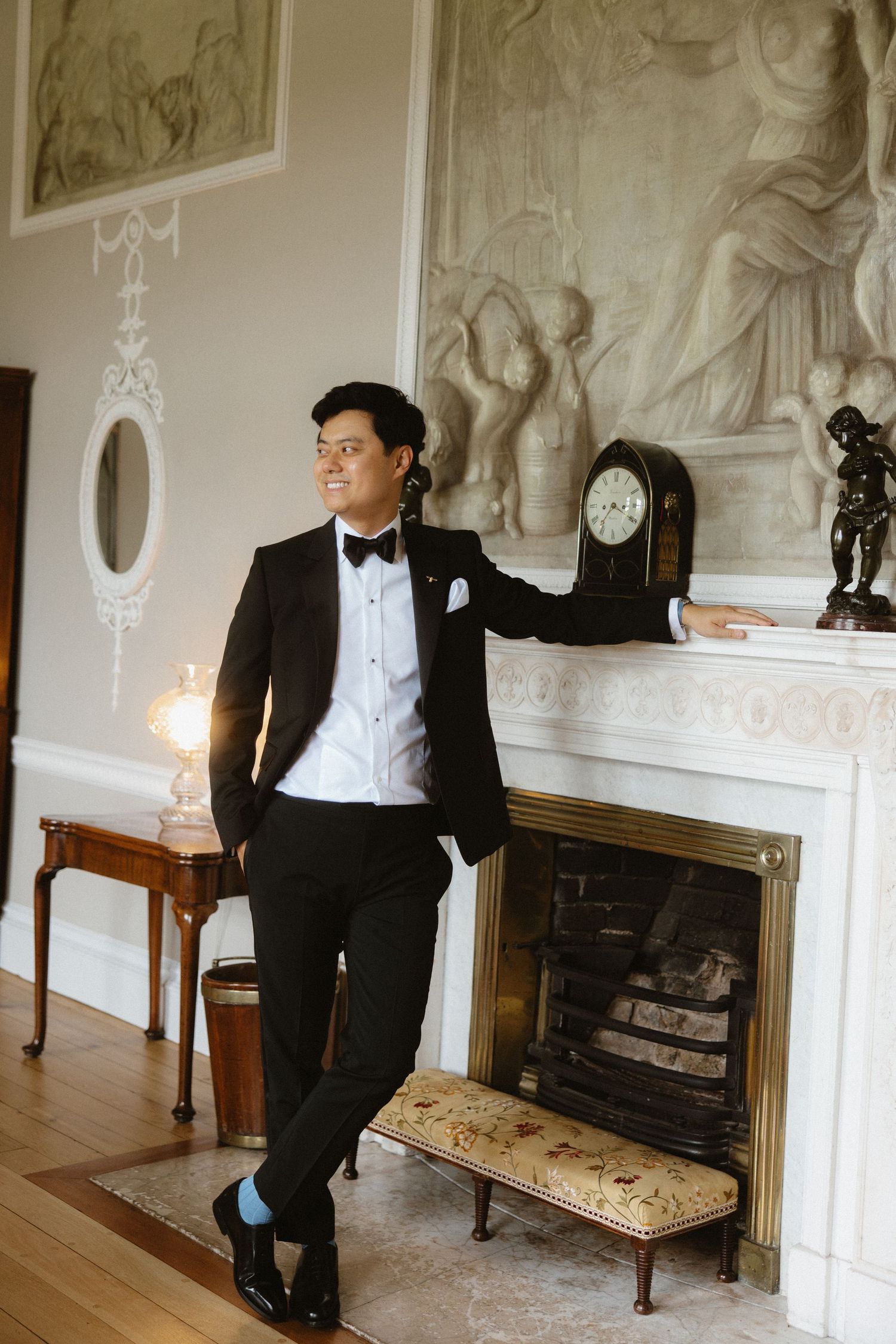 Formal portrait of someone in a tuxedo leaning against an ornate fireplace mantle.