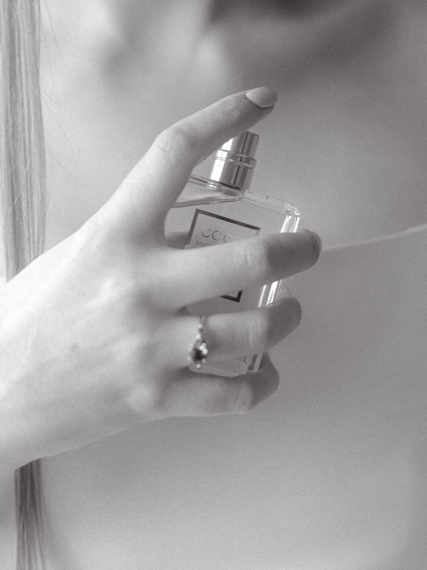 Close-up black and white photo of hand holding perfume bottle with engagement ring visible.