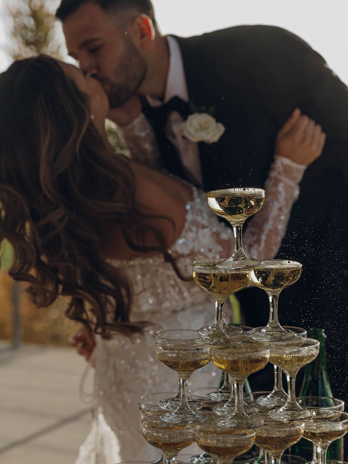 A champagne tower of coupe glasses stands elegantly in the foreground of a romantic wedding celebration moment.