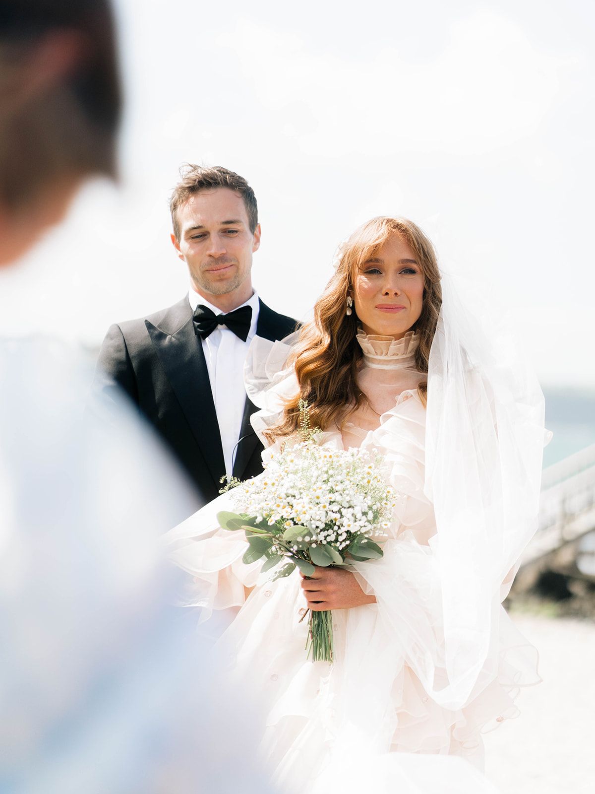A couple in wedding attire stands together during their ceremony holding a white flower bouquet.
