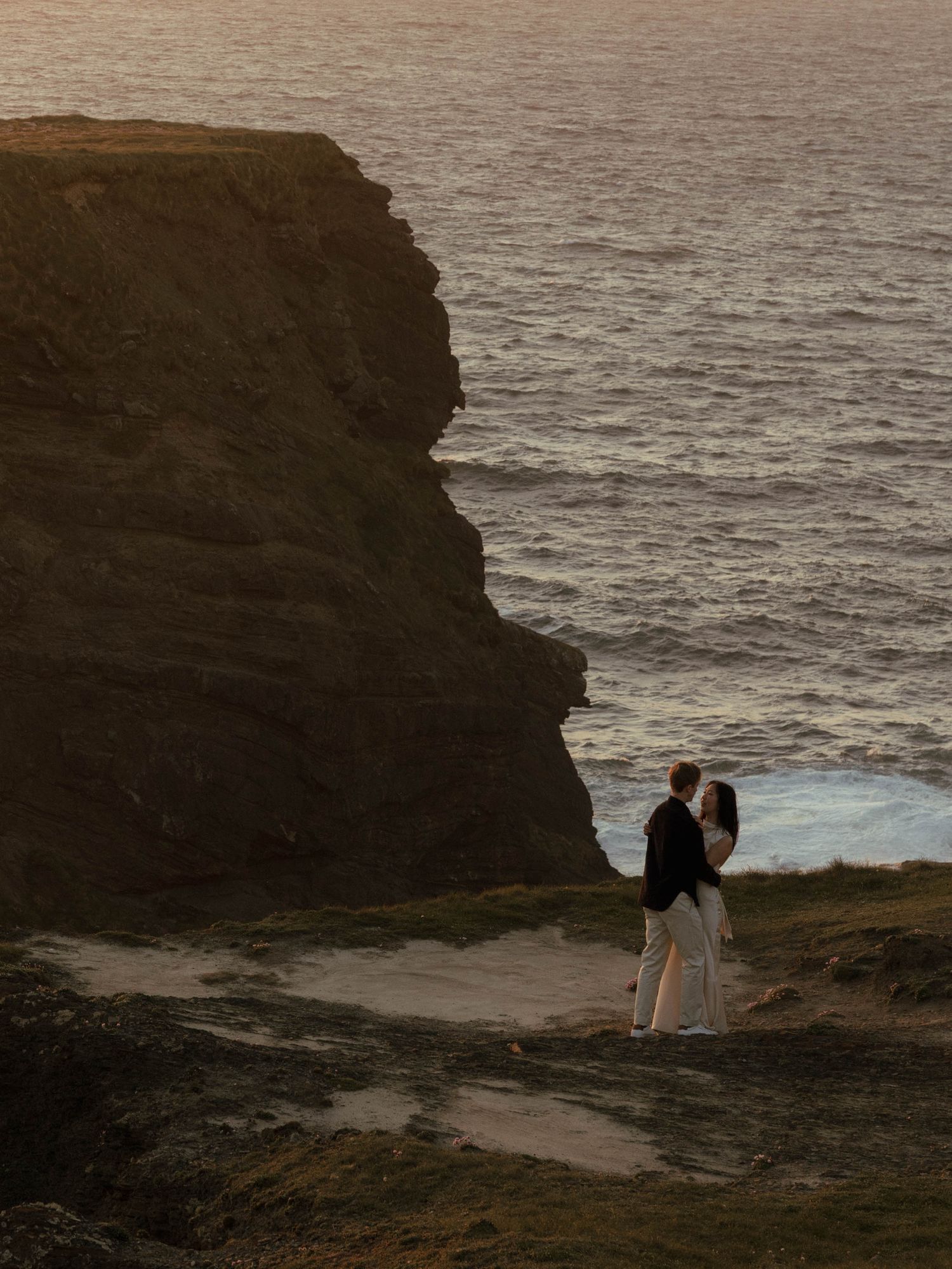 A couple standing on dramatic coastal cliffs overlooking the ocean at sunset.
