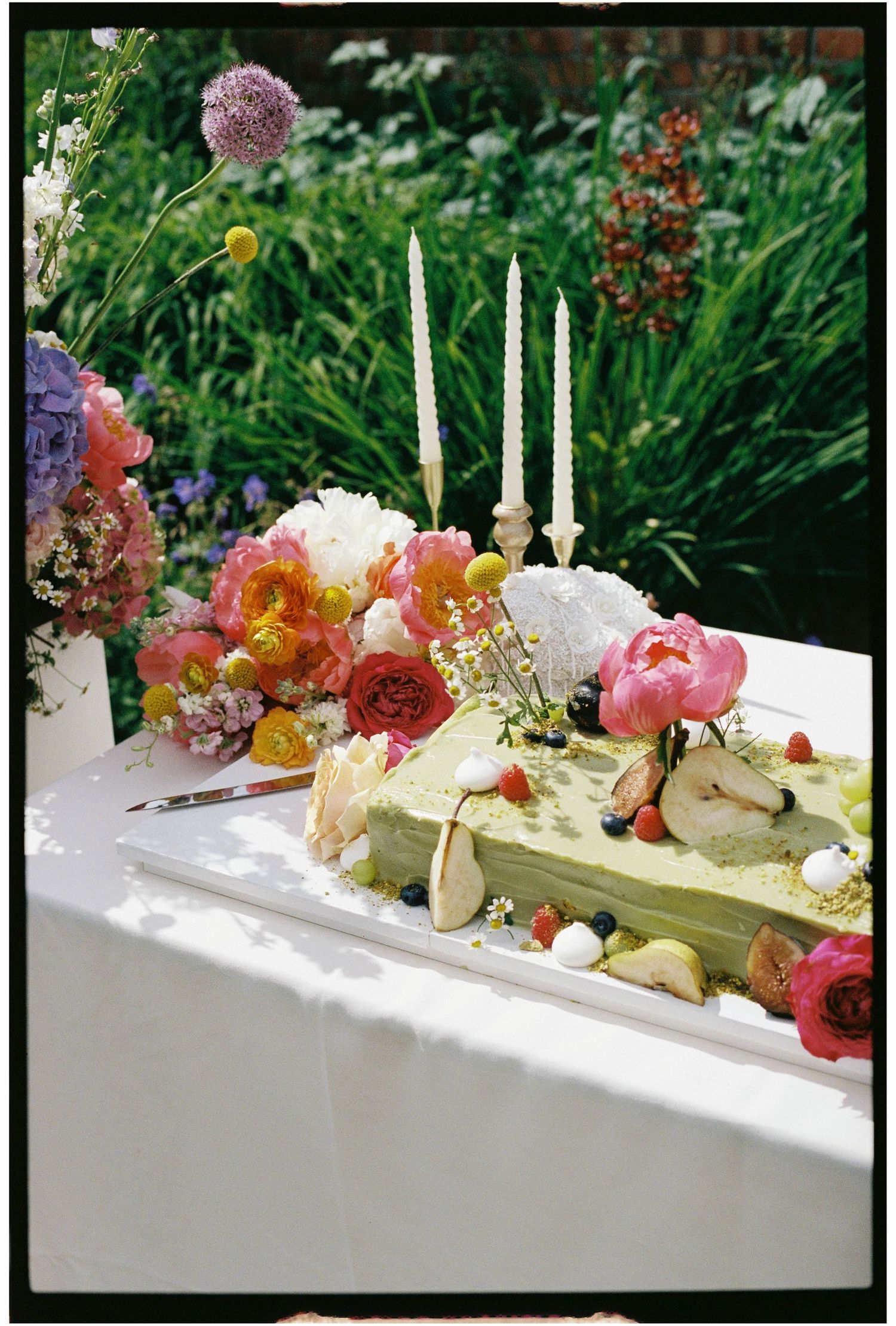 Green wedding cake decorated with fresh flowers, berries, and candles on white outdoor table.