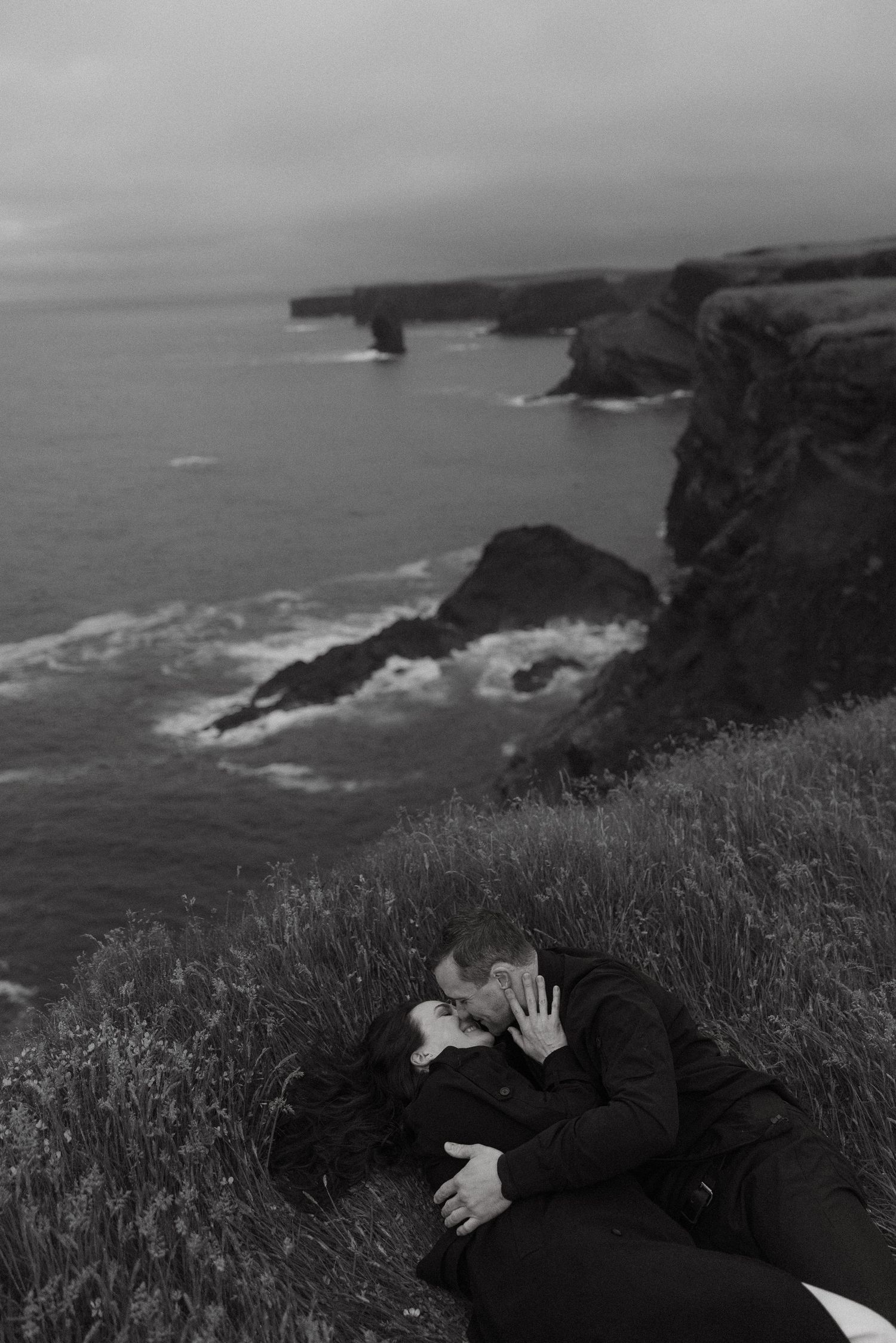 Black and white photo of a couple kissing on a grassy cliff overlooking dramatic ocean coastline.