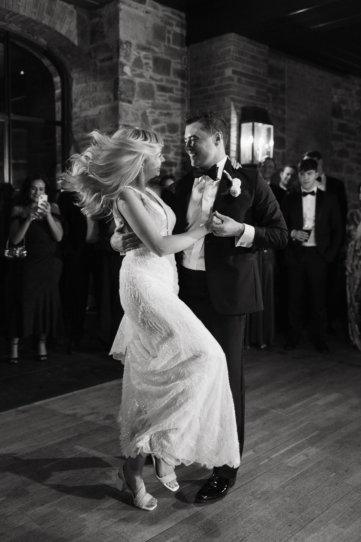 A couple dances energetically at a wedding reception in a rustic brick-walled venue with a wooden floor.