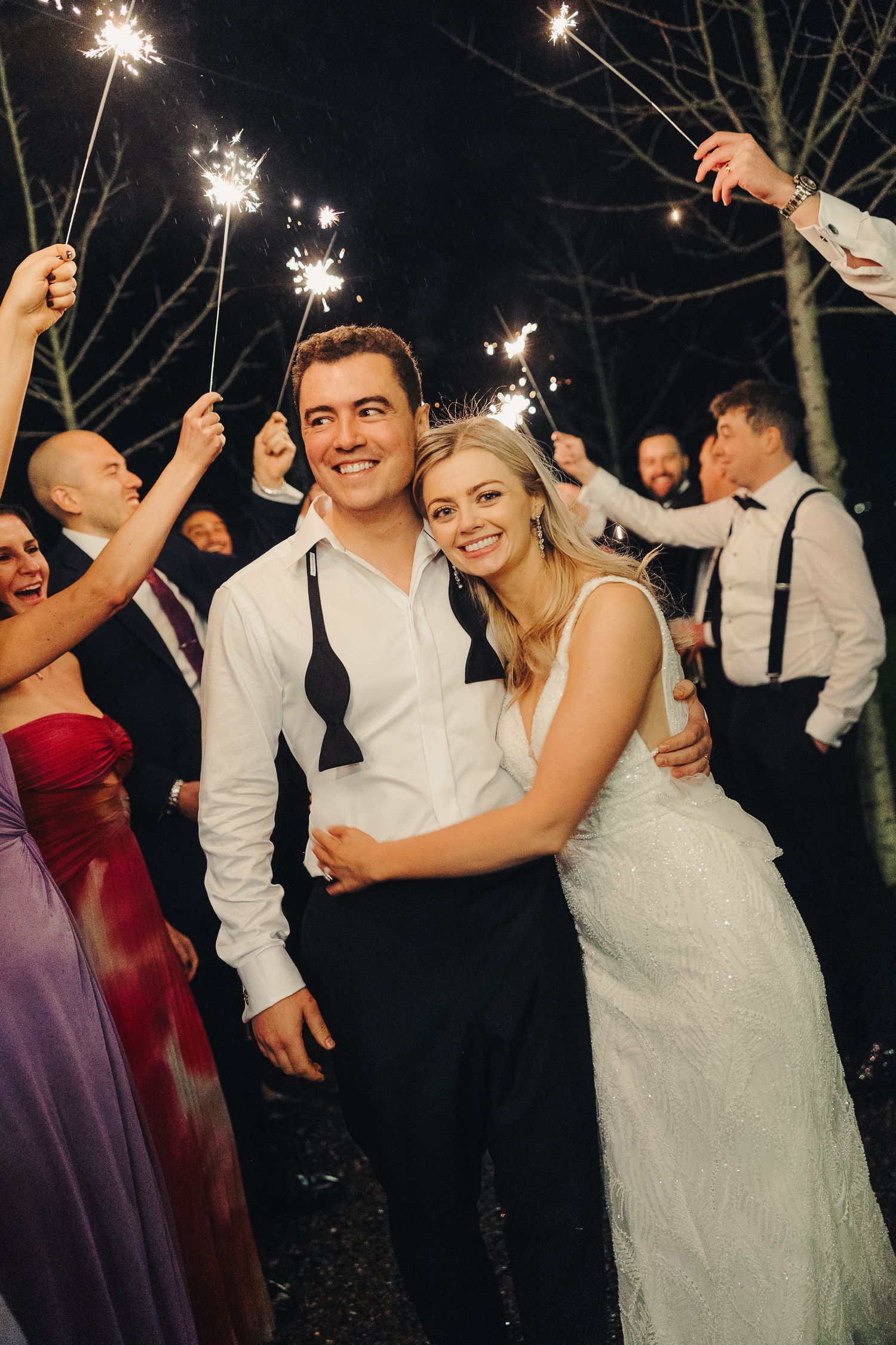 Wedding celebration at night with sparklers glowing as guests celebrate around a couple in formal attire.