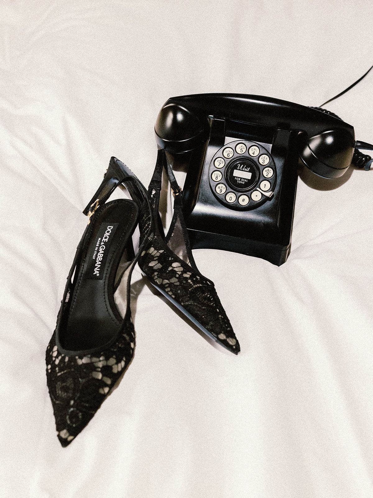 Vintage black rotary telephone displayed alongside black studded high heel shoes on white fabric.