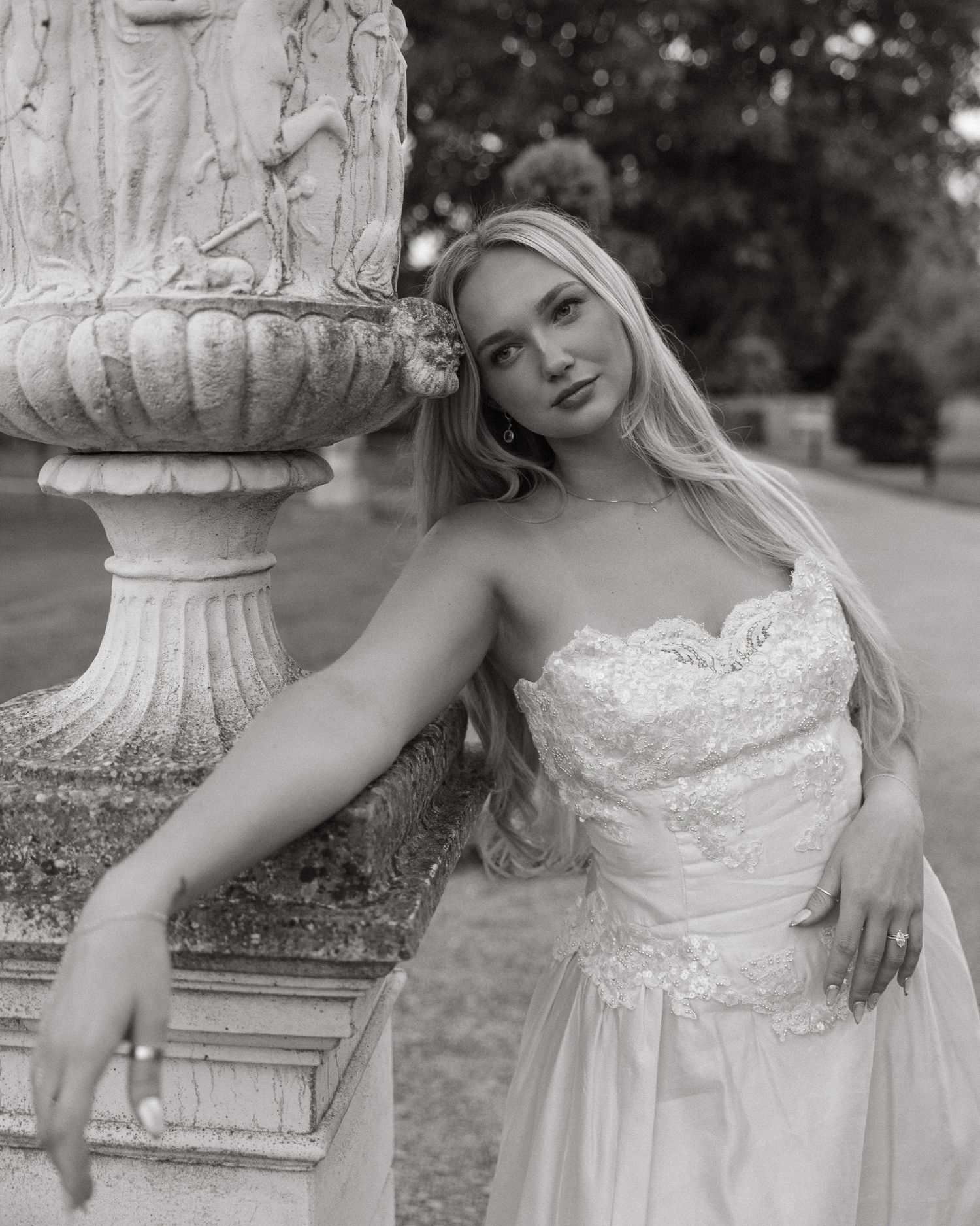 Black and white photo of a blonde woman in a strapless lace gown leaning against an ornate stone fountain.