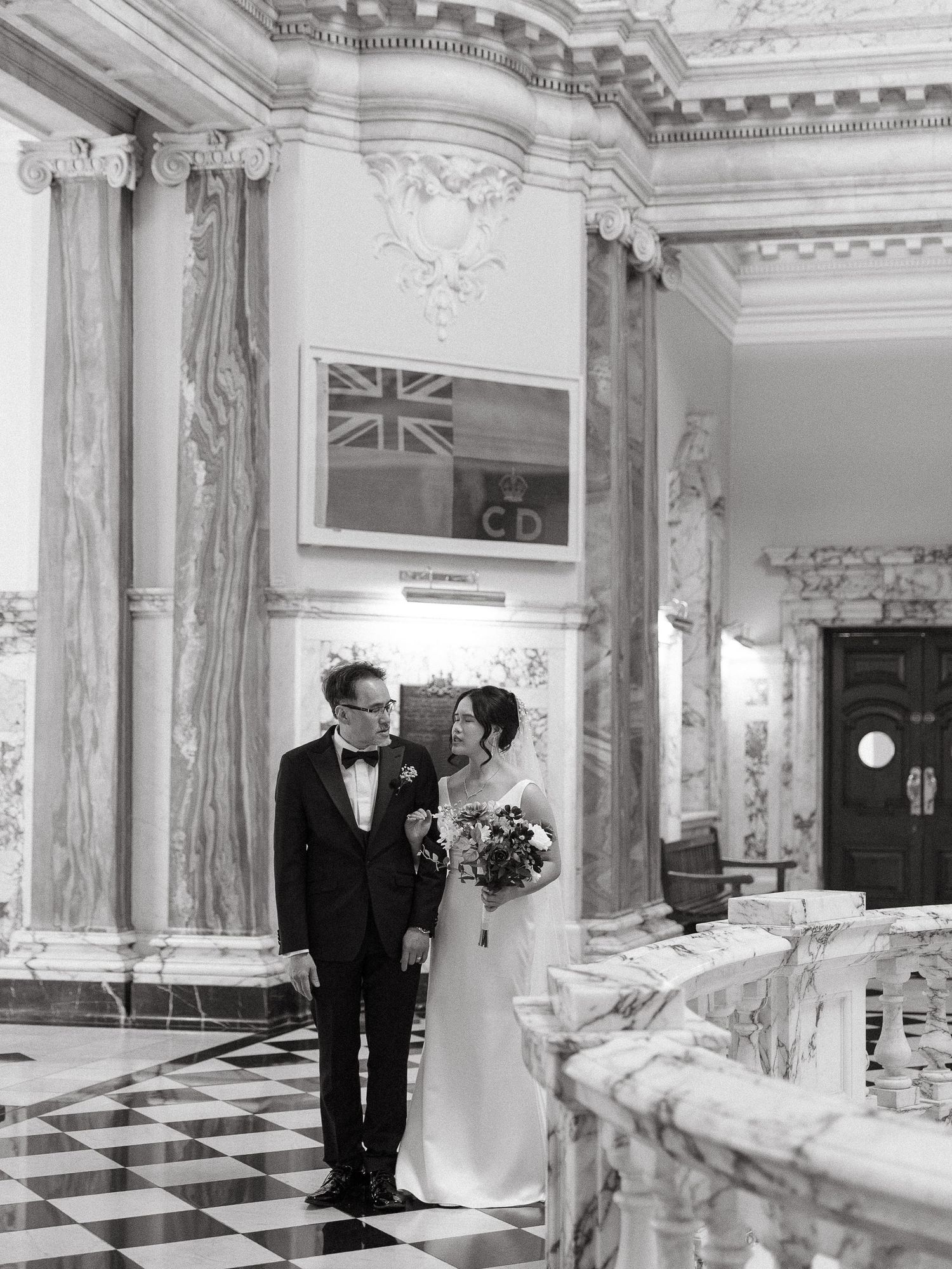 A black and white wedding photo in an elegant marble interior with ornate columns and a British flag on display.