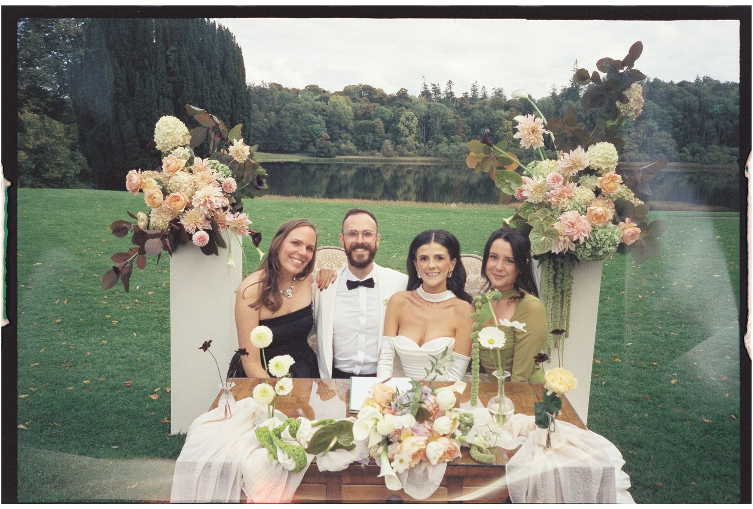 A wedding party poses in a lush garden setting with peach and blush floral arrangements.