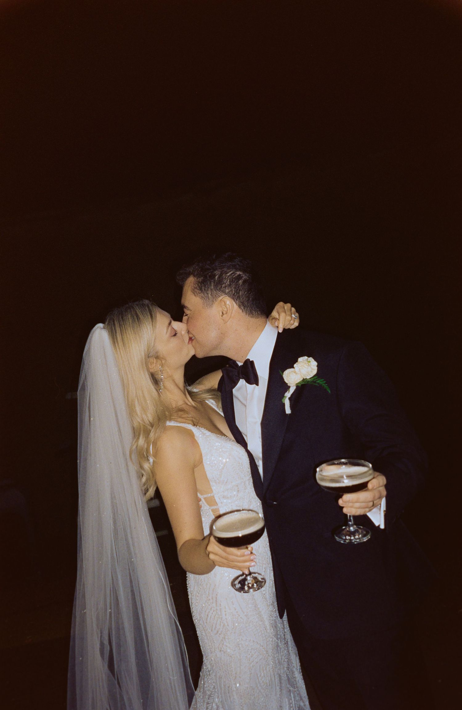 A couple shares a romantic kiss at their wedding reception while holding cocktail glasses against a dark background.