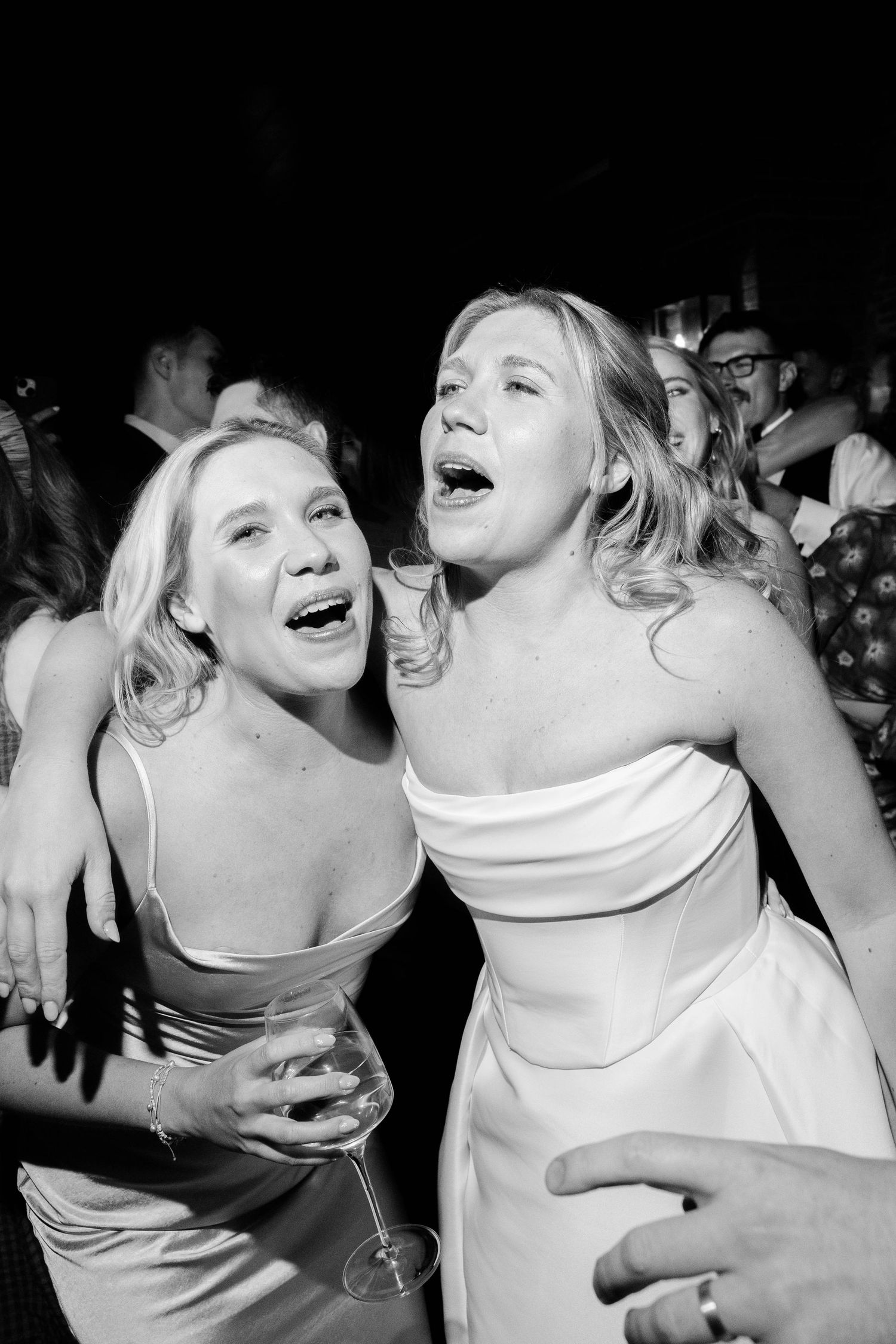 Two women in elegant dresses laugh together during a wedding reception celebration.