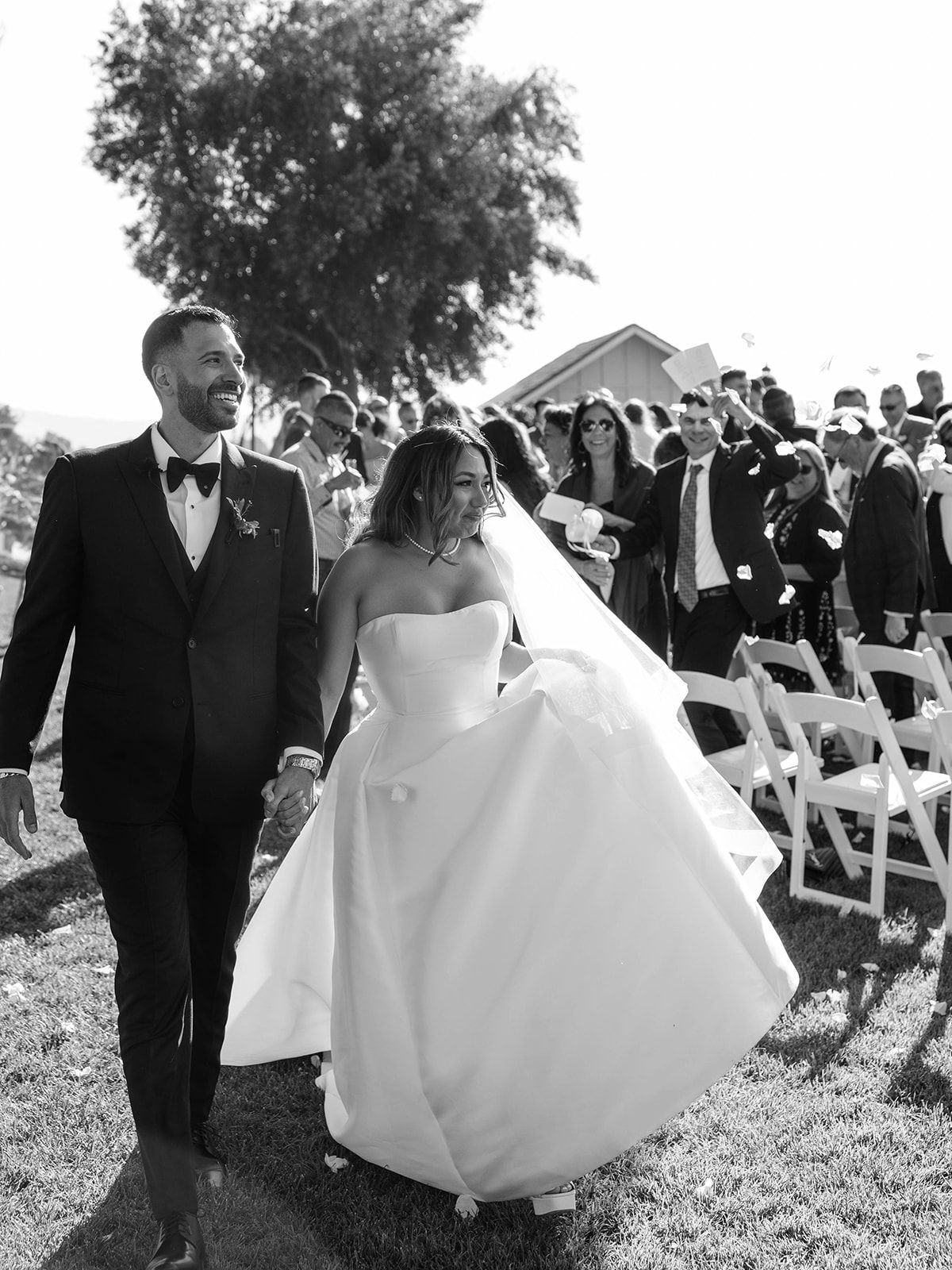 A couple in wedding attire walks together on grass near white chairs at an outdoor ceremony venue.