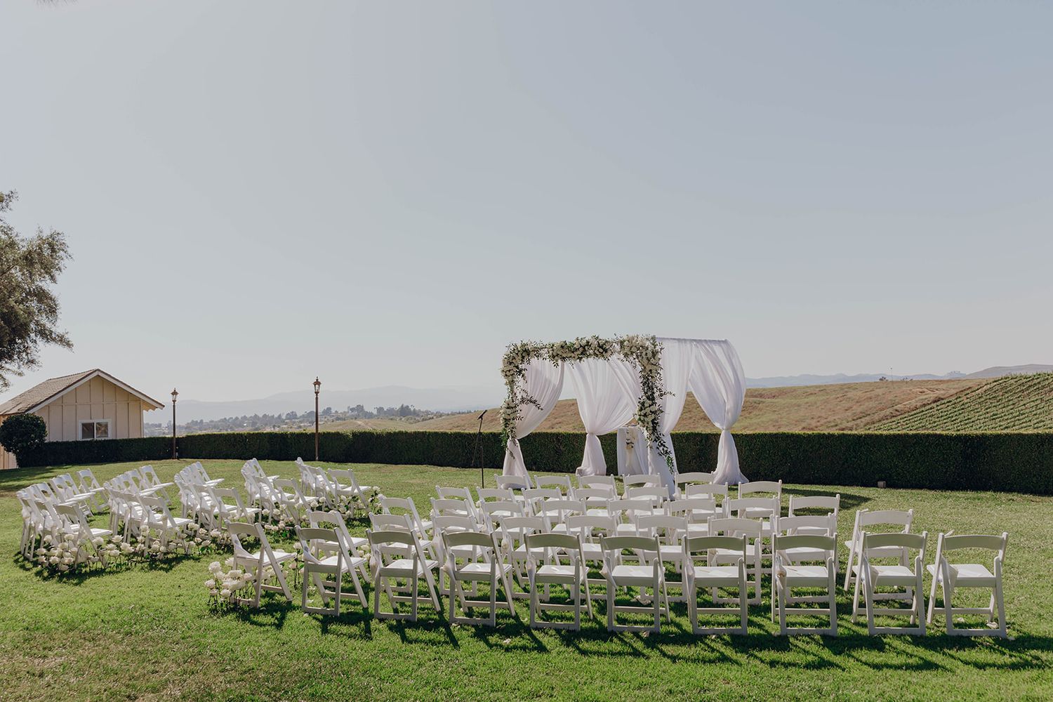 White chairs arranged in rows facing an elegant wedding arch setup on a grassy hillside under an overcast sky.