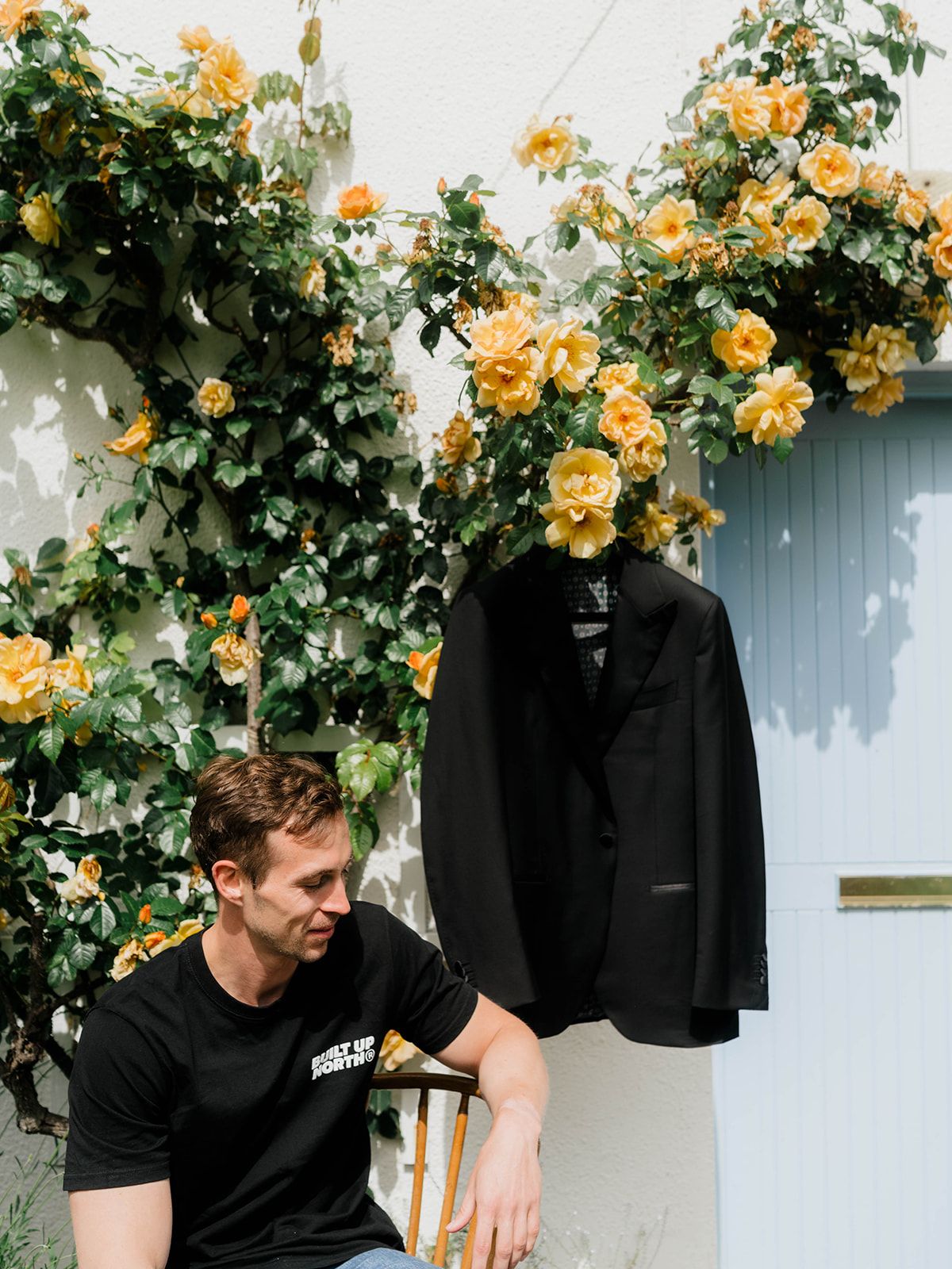 Black suit jacket displayed against wall with blooming yellow climbing roses and seated figure.