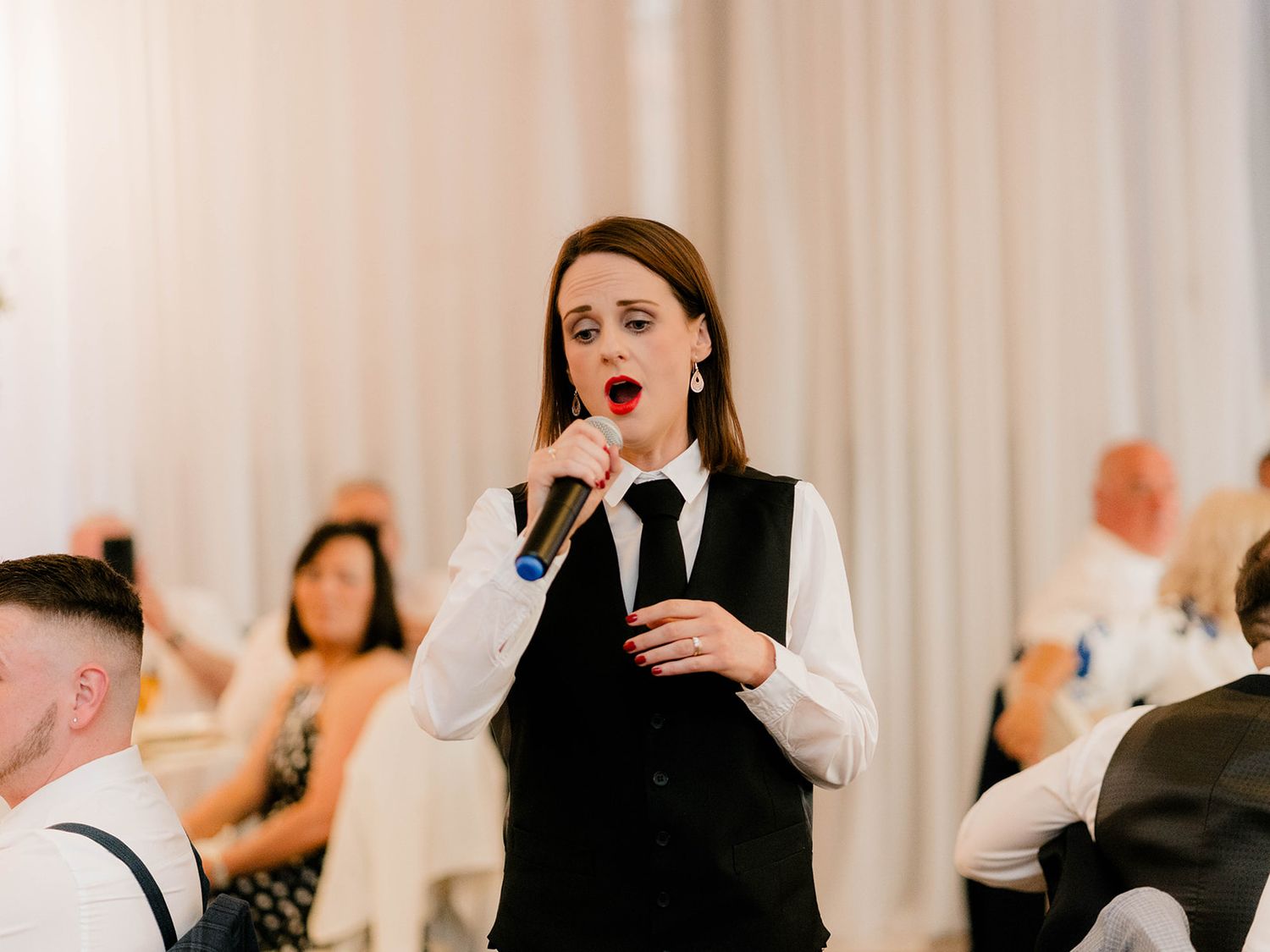 A singer in formal black vest and white shirt performs emotionally at an event with guests visible in the background.