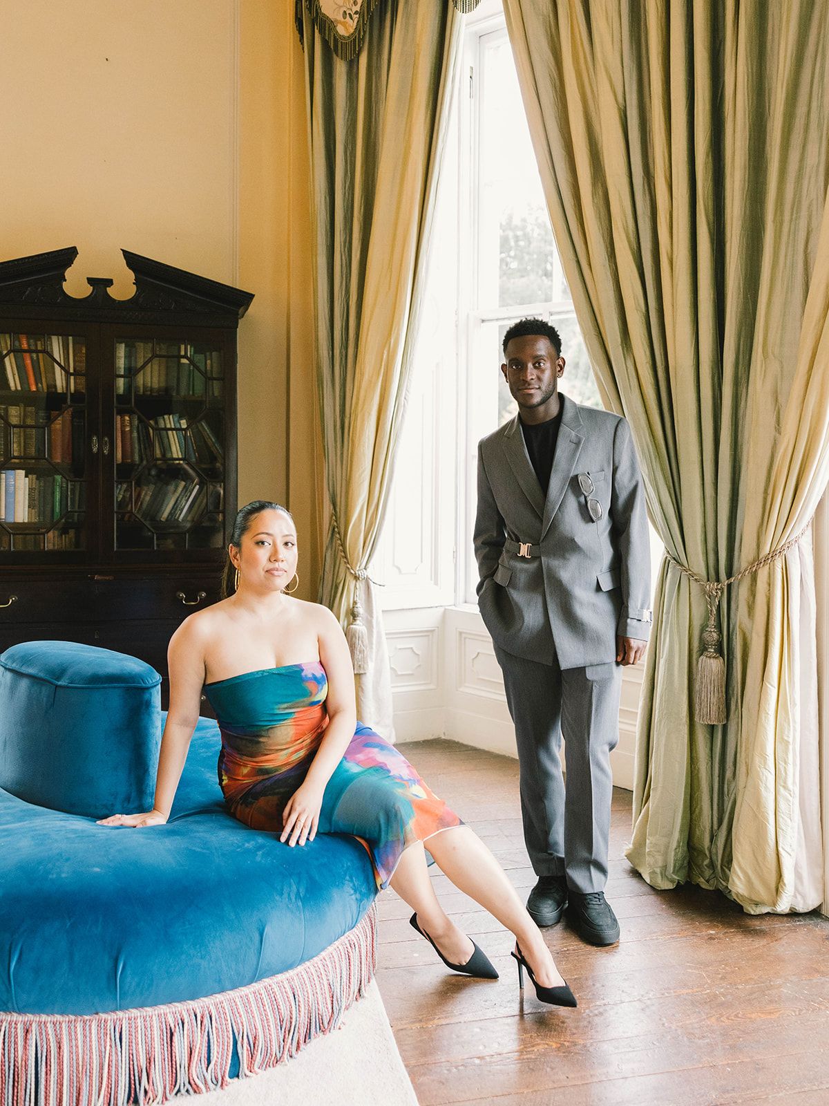 A fashionable couple poses in an elegant room with a blue velvet chaise lounge and floor-length green curtains.