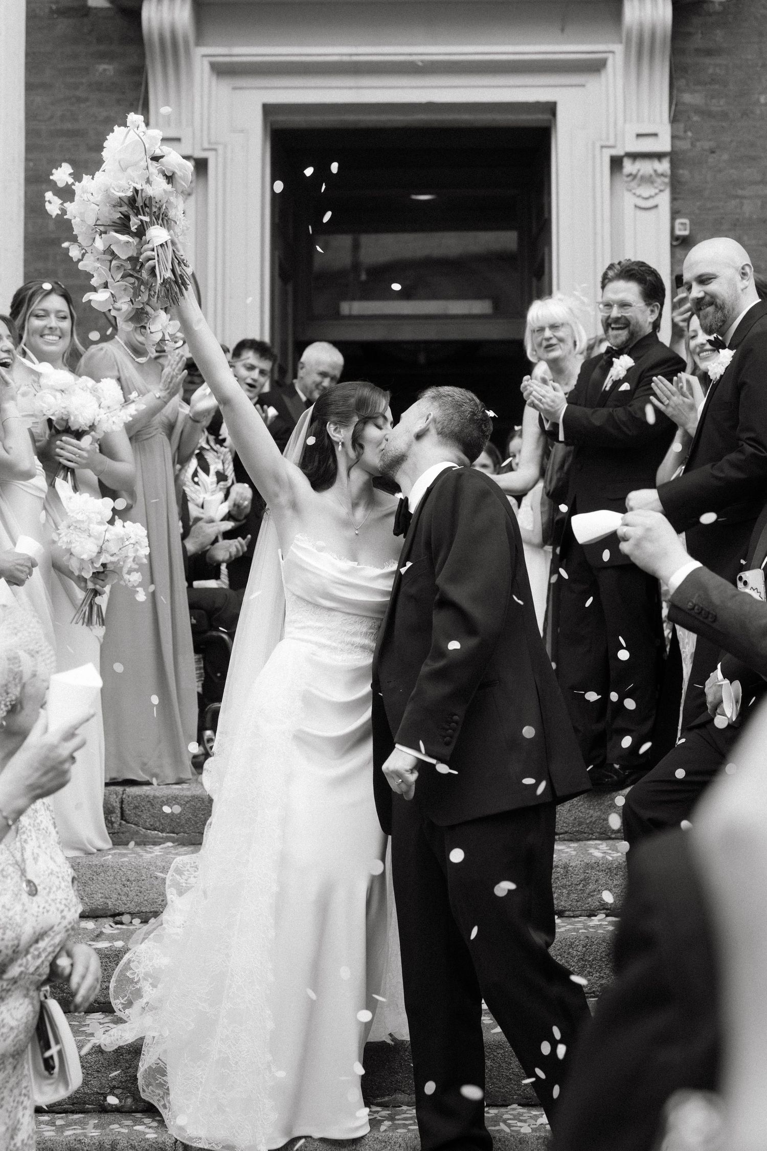 Black and white photo of bride and groom kissing on church steps while guests celebrate with confetti.