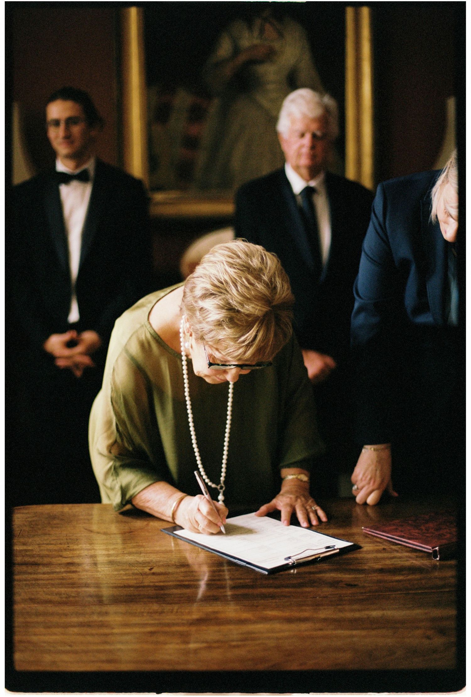 A woman in a green dress signs a wedding register document at a table while guests watch in a historic room.