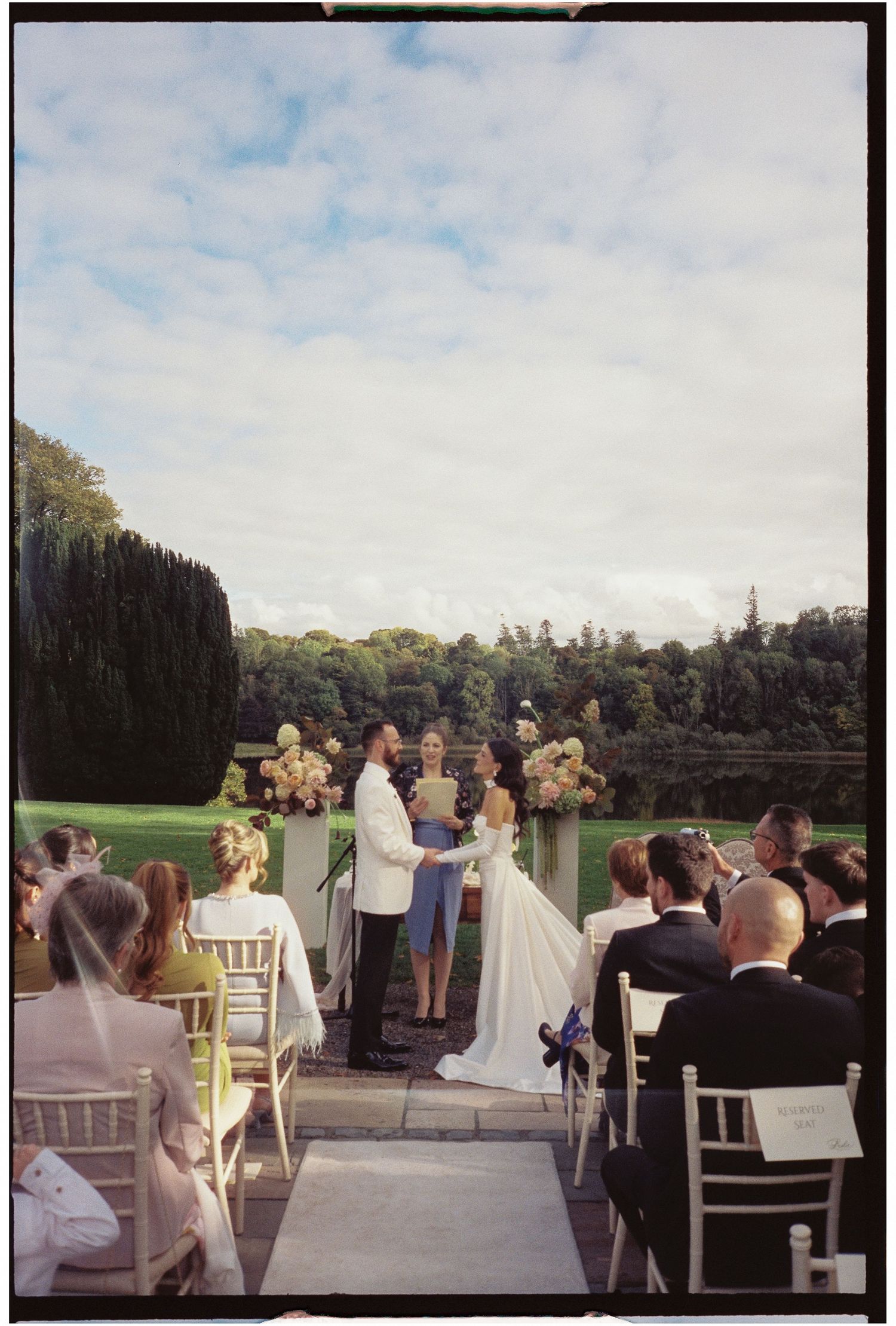 Outdoor wedding ceremony with guests seated in white chairs overlooking a scenic landscape on a cloudy day.