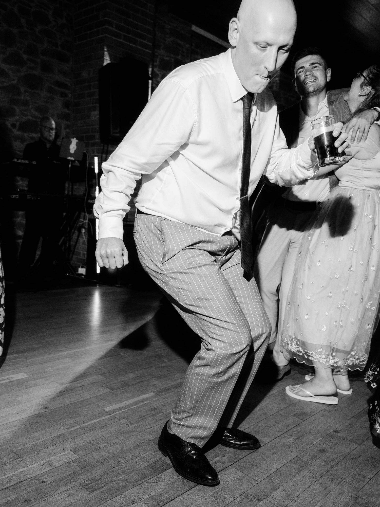 A person in striped pants and white shirt dances energetically on a wooden dance floor while others watch from the side.