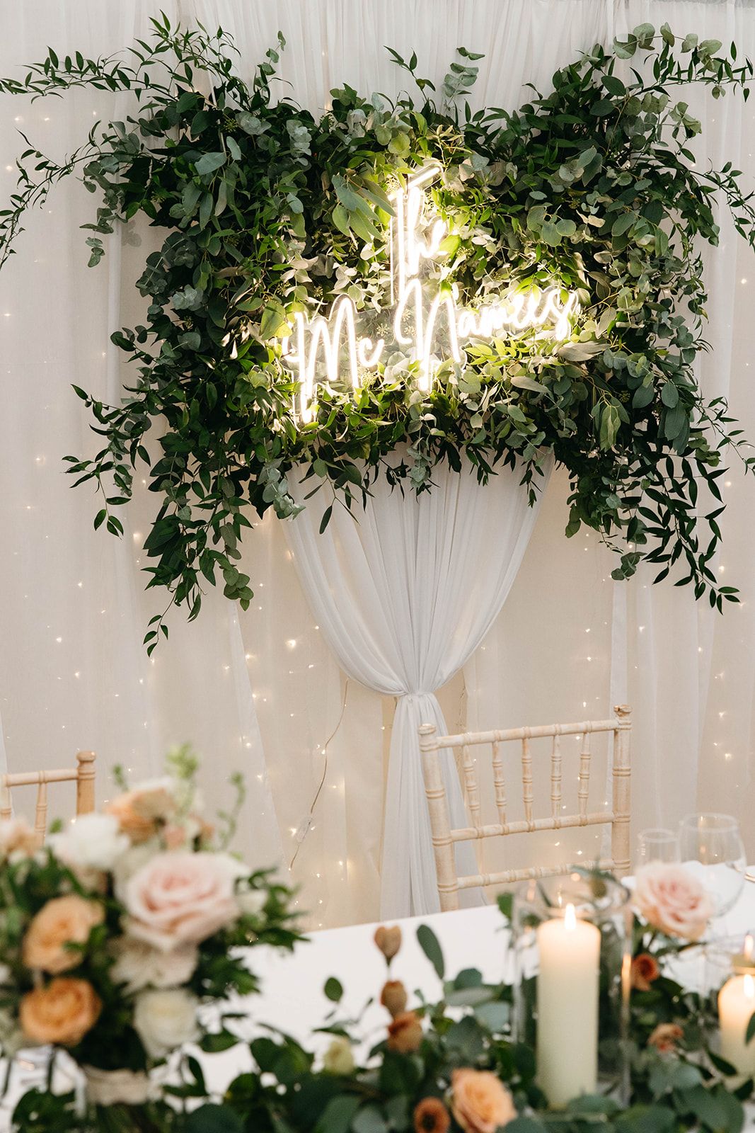 Elegant wedding decor featuring a neon 'Mr & Mrs' sign surrounded by lush greenery and soft lighting with white drapes.