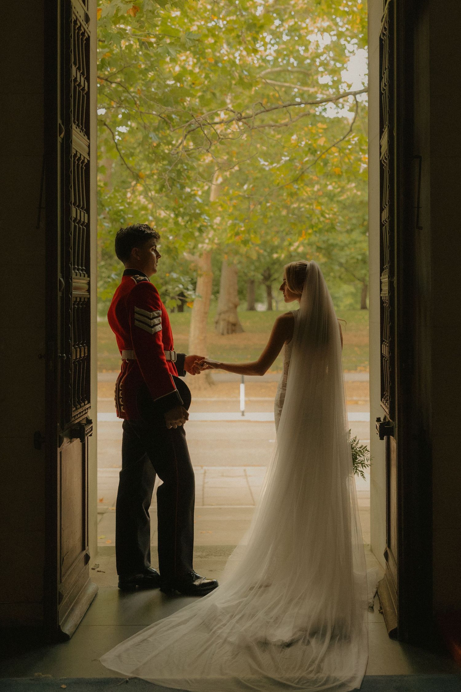 A silhouetted couple in wedding attire stands in a doorway framed by green trees on a bright autumn day.