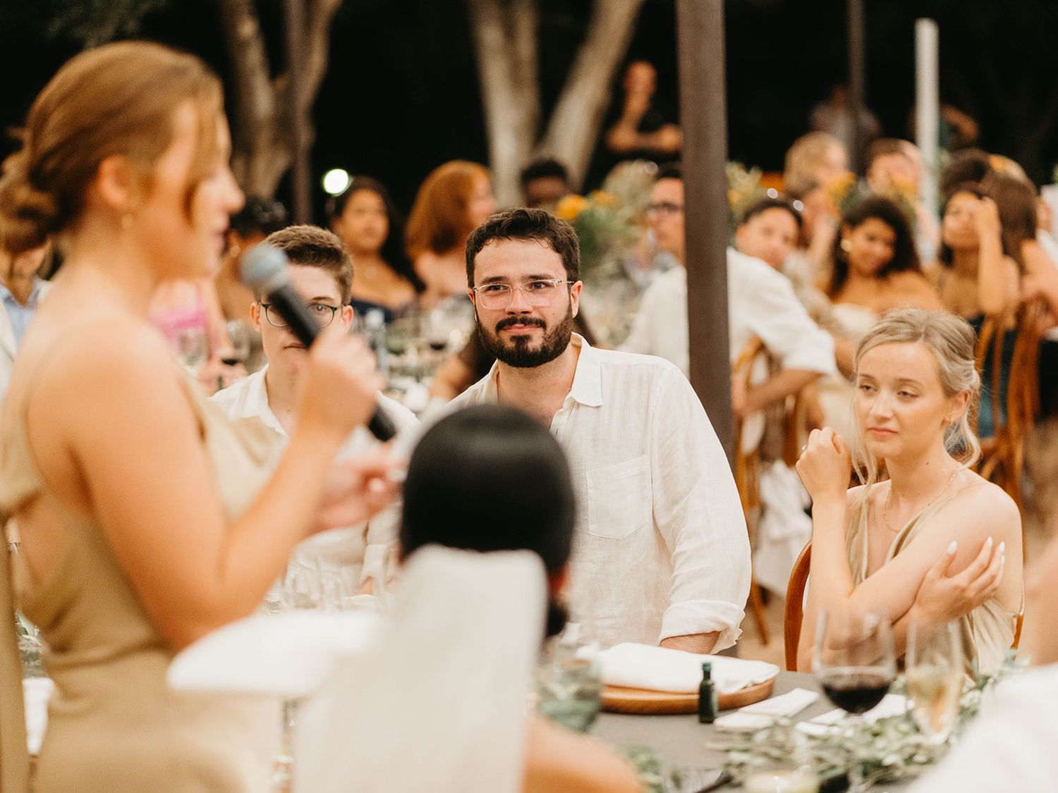 Wedding guests react with joy during an outdoor reception dinner surrounded by trees and evening light.