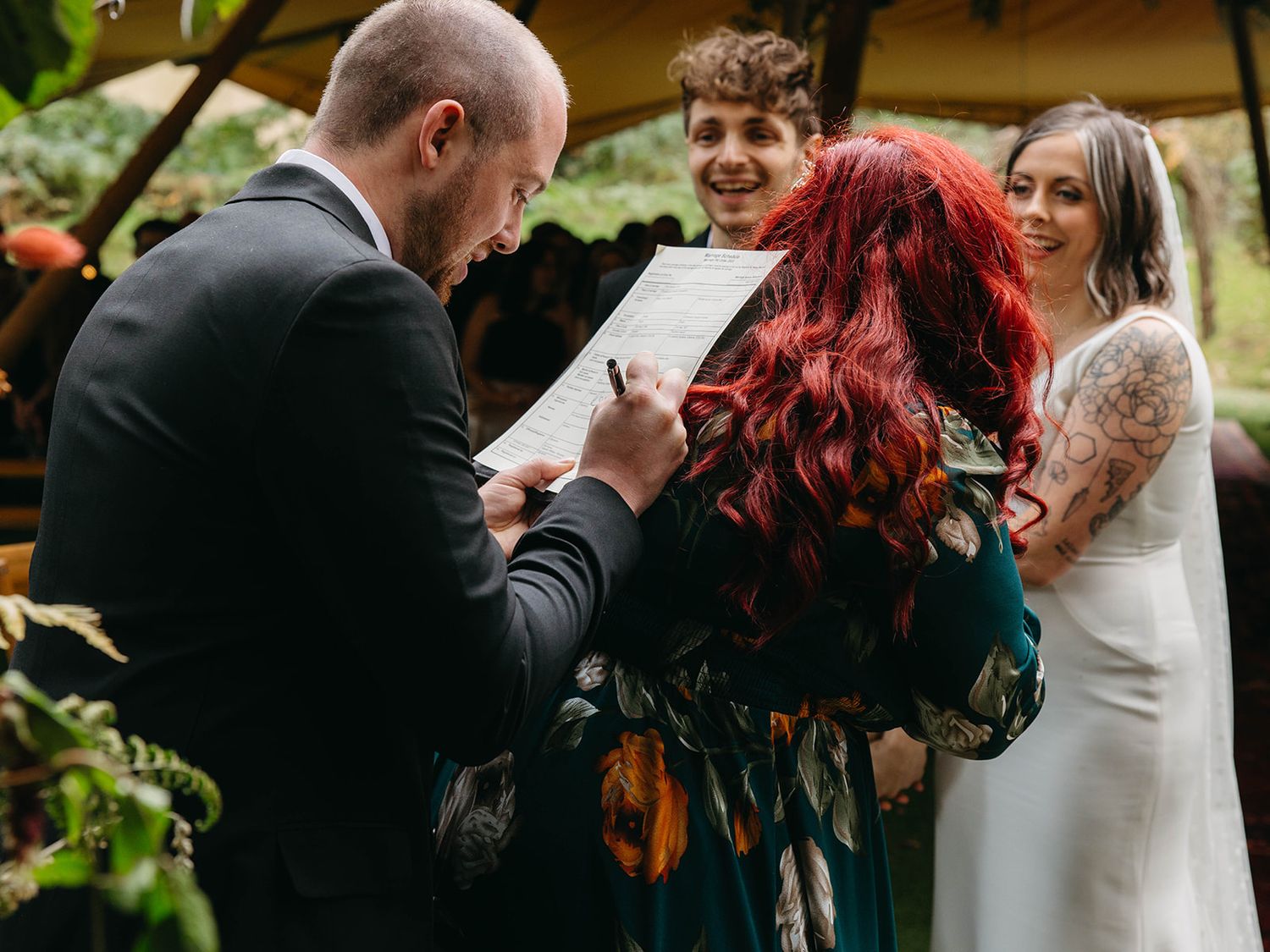 An intimate outdoor wedding ceremony with an officiant reading from papers while attendees gather under greenery.